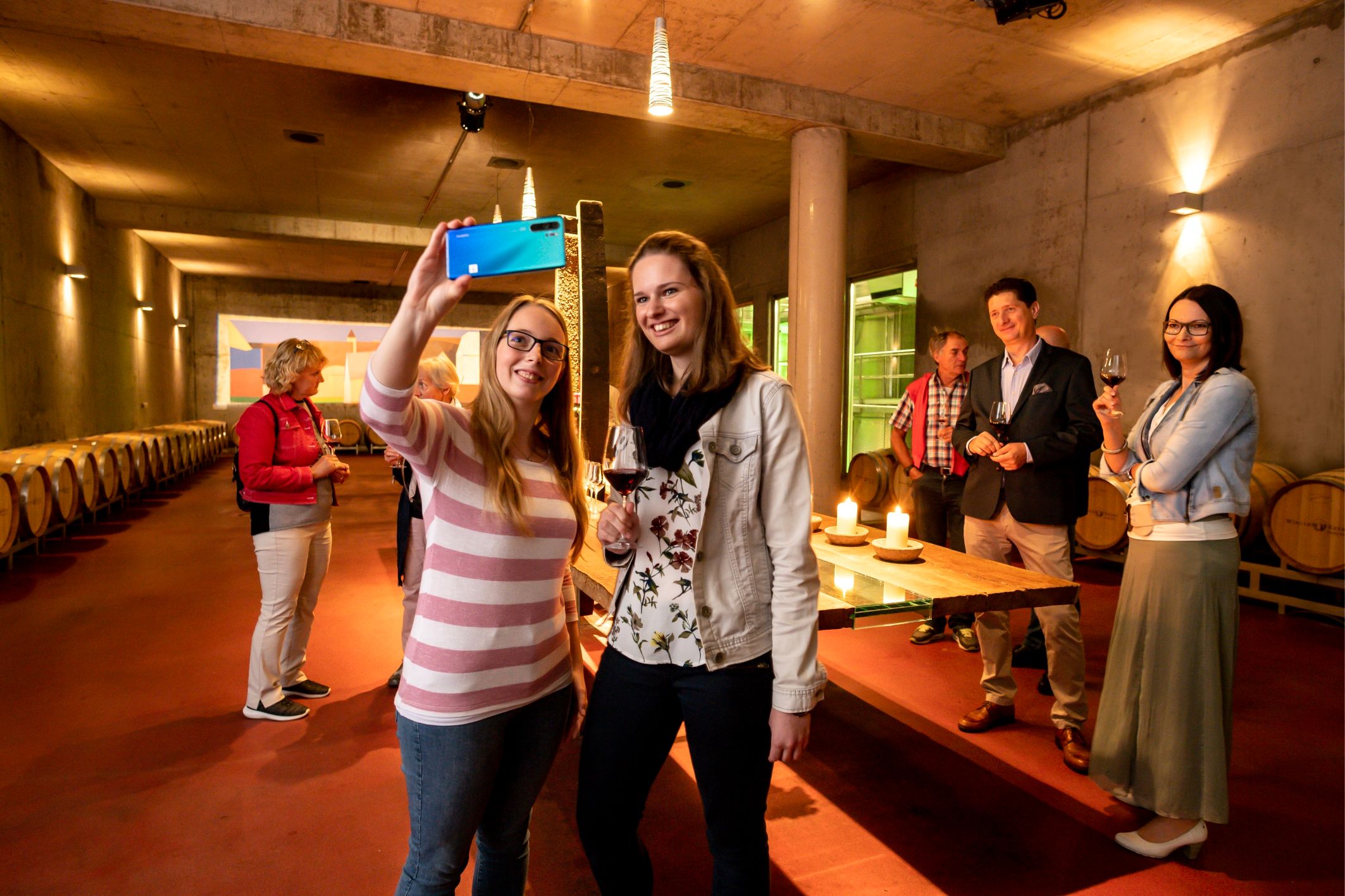 Junge Menschen stehen im Barriquekeller der Winzer Krems, wobei 2 Mädchen ein Handy hochhalten und fotografieren.