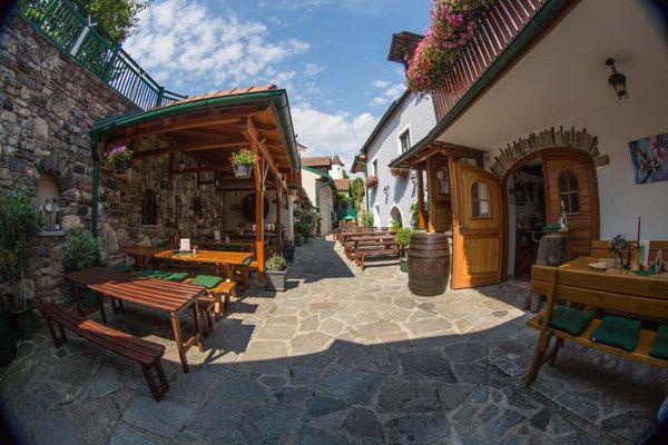 Ein gemütlicher Innenhof mit Holzbänken, Tischen und Blumen. Steinboden und überdachte Sitzbereiche. Sonniges Wetter mit blauem Himmel.