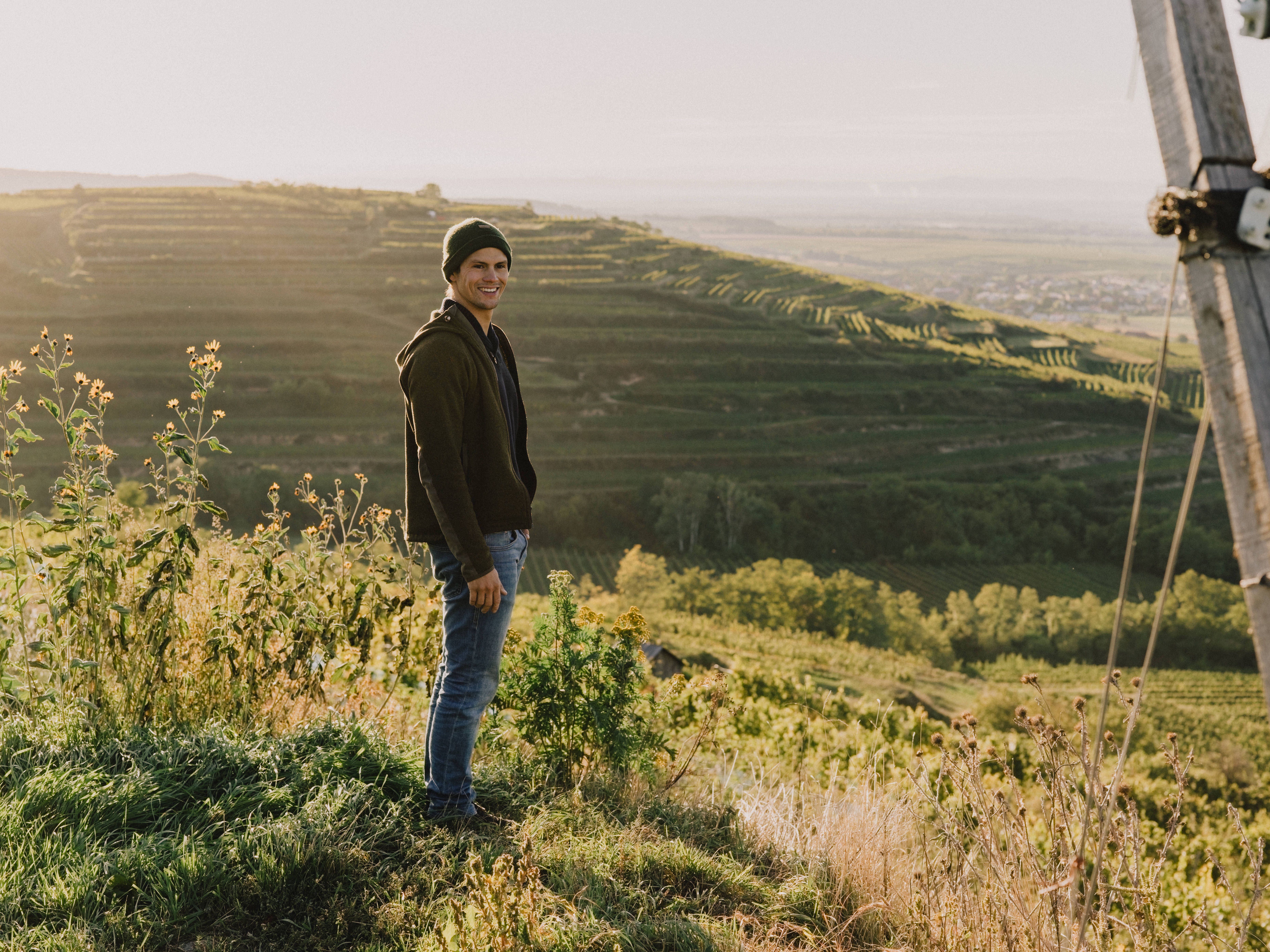 In der sanften Morgenröte erstrahlen die Weinberge in voller Pracht. Ein junger Mann genießt die atemberaubende Aussicht über die sanften Hügel, während die ersten Sonnenstrahlen die Landschaft in goldenes Licht tauchen. Diese Idylle lädt dazu ein, die Seele baumeln zu lassen und die Schönheit der Natur zu erleben.