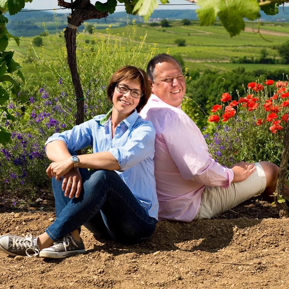 Two people sitting back to back in a vineyard with flowers in the background.