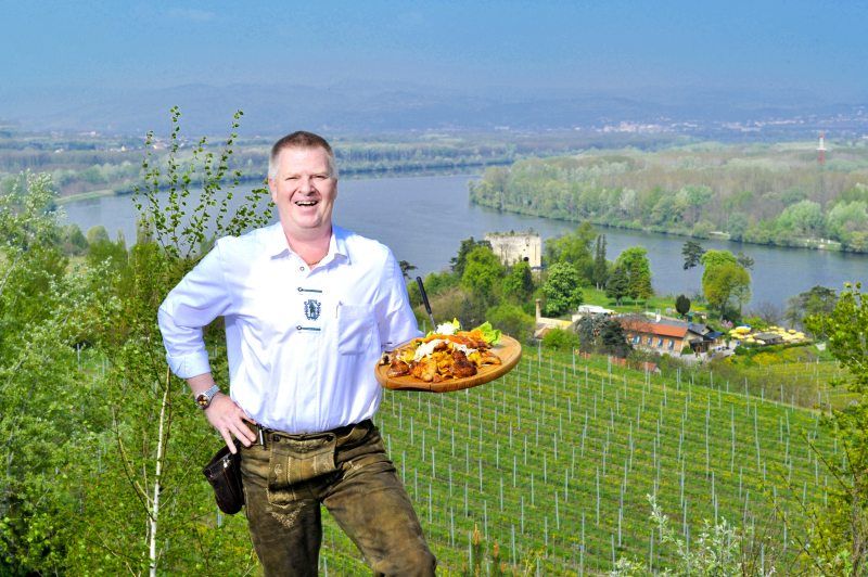 Ein Mann in traditioneller Kleidung hält ein Tablett mit Essen vor einer Landschaft mit Fluss und Weinbergen.