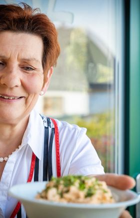 Eine freundliche Gastgeberin präsentiert stolz eine köstliche Jause mit Liptauer, die zum Verweilen und Genießen einlädt. Die warme Atmosphäre und die frischen Zutaten versprechen ein unvergessliches Geschmackserlebnis inmitten der Natur.