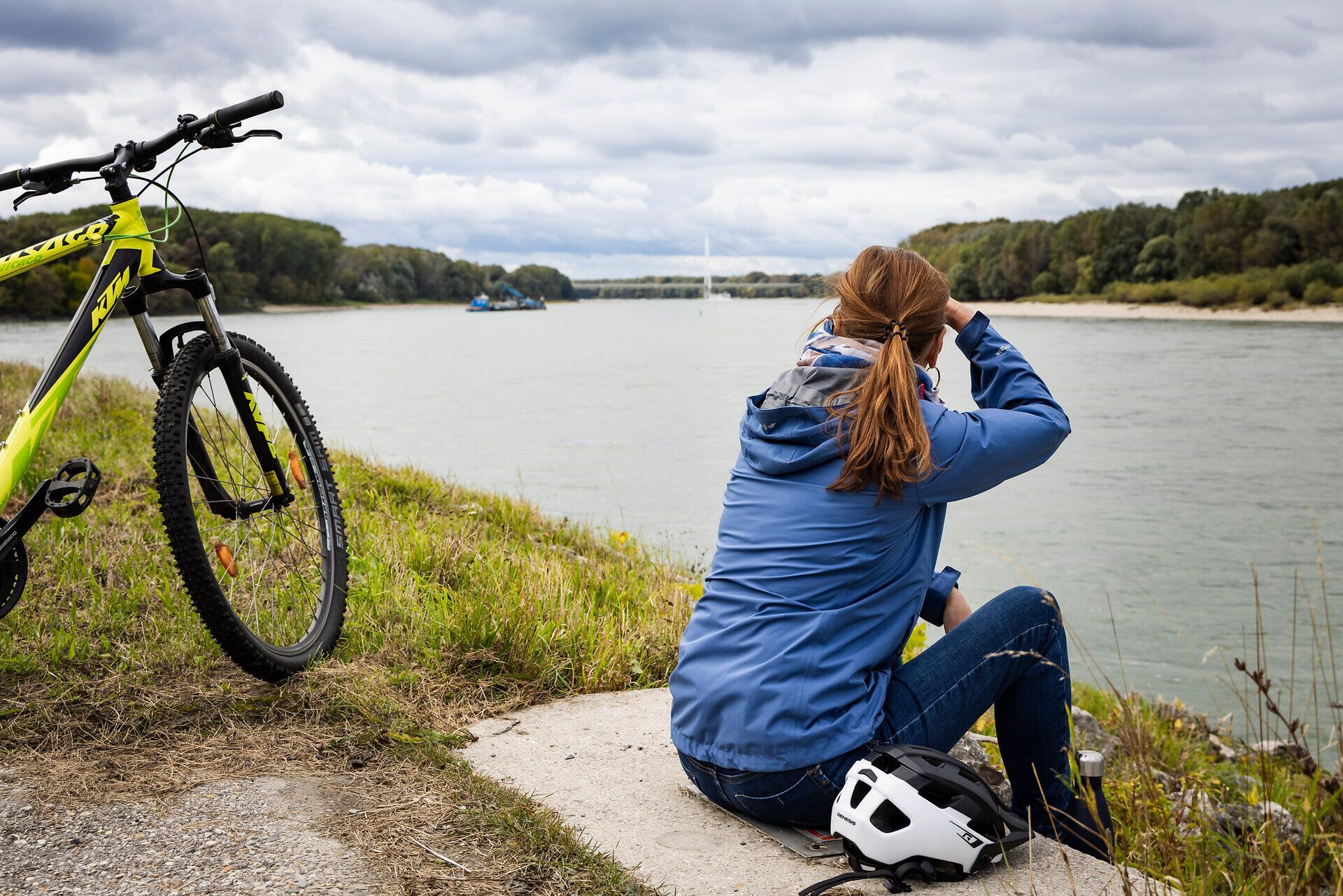 Frau sitzt am Donauufer und blickt aufs Wasser. Neben ihr steht ein Fahrrad.