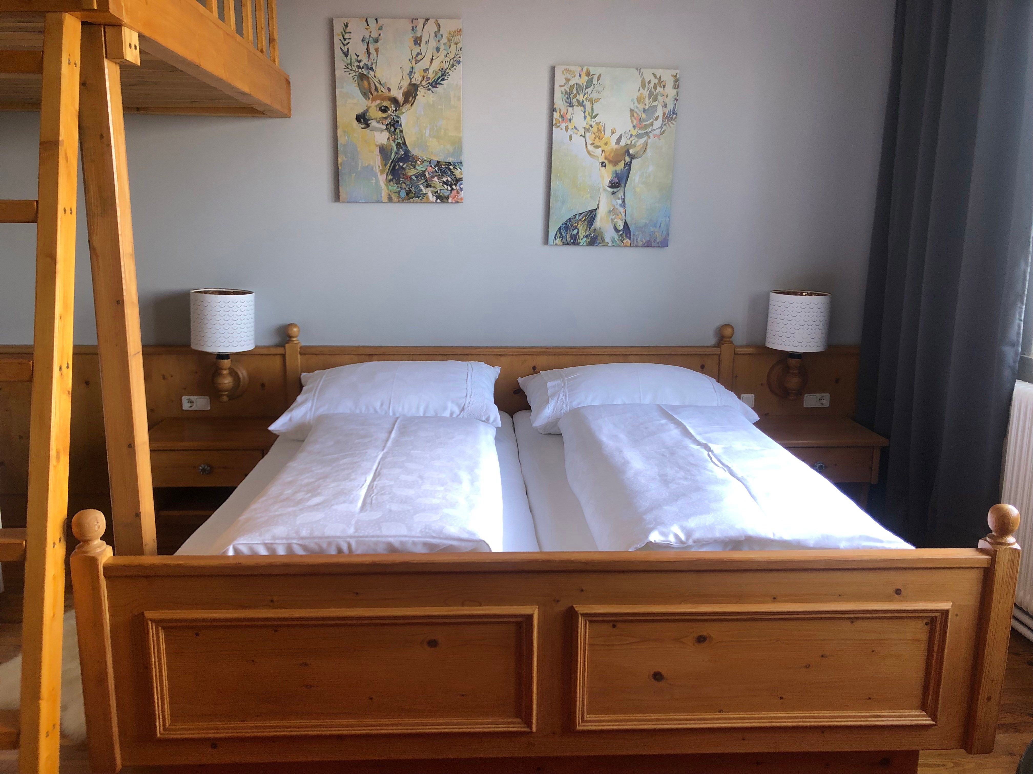 Gemütliches Schlafzimmer mit Holzmöbeln, Doppelbett, zwei Hirschbildern an der Wand und einer Leiter zu einem Hochbett.