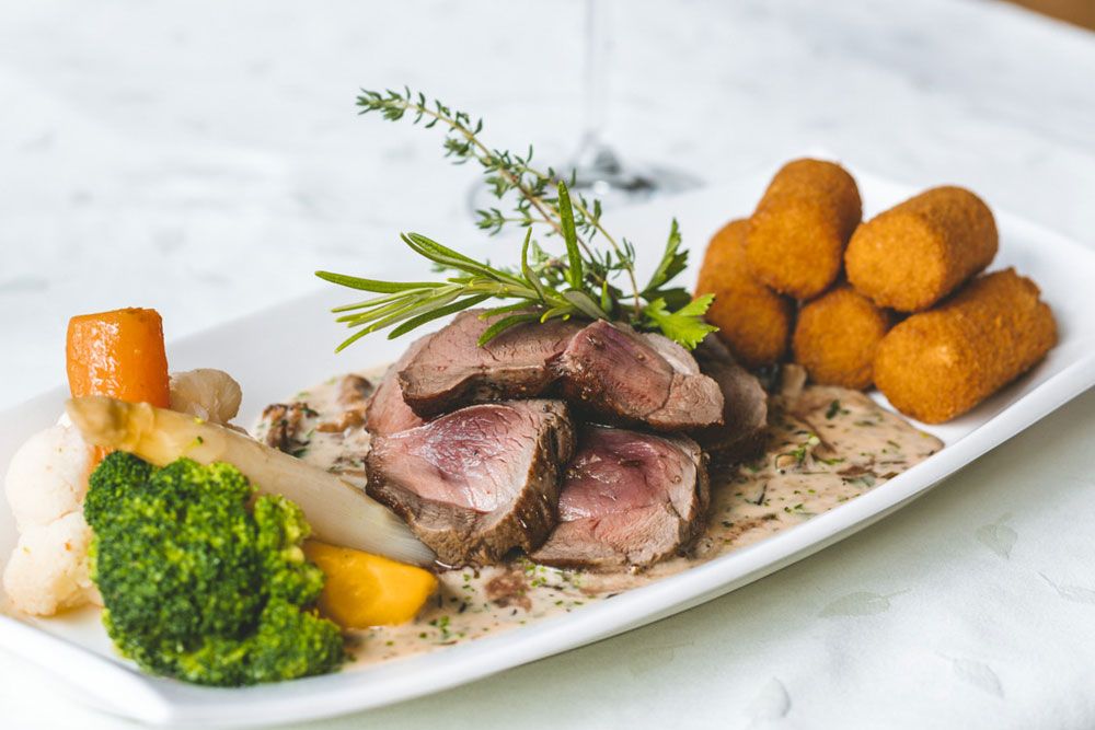 Gourmetgericht mit Fleisch, Gemüse und Kroketten auf weißem Teller.