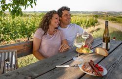Paar sitzt entspannt an einem Holztisch im Weingarten mit Weinflaschen.
