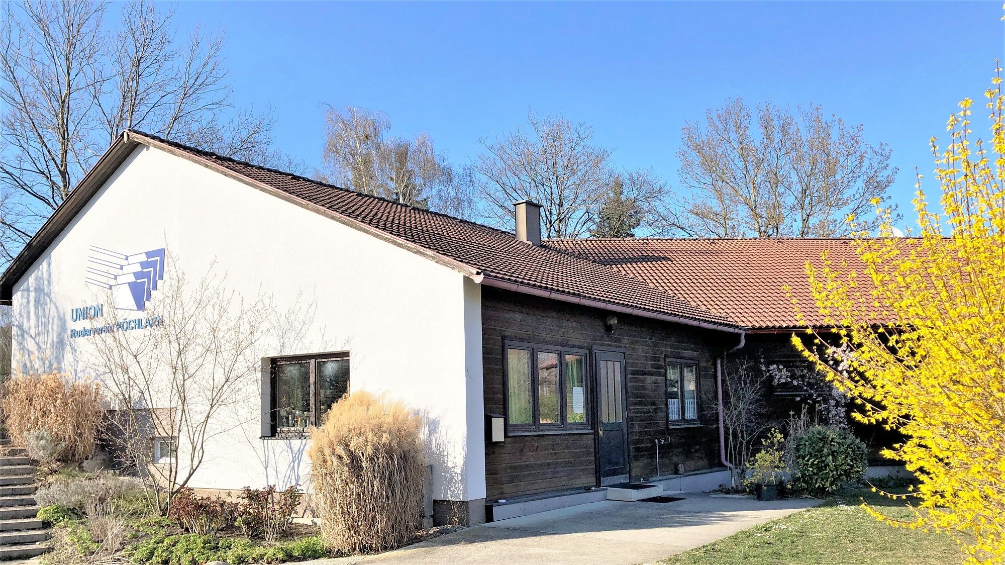 Bootshaus des Union Rudervereins Pöchlarn mit weißer Fassade und braunem Dach, umgeben von Bäumen und Sträuchern.