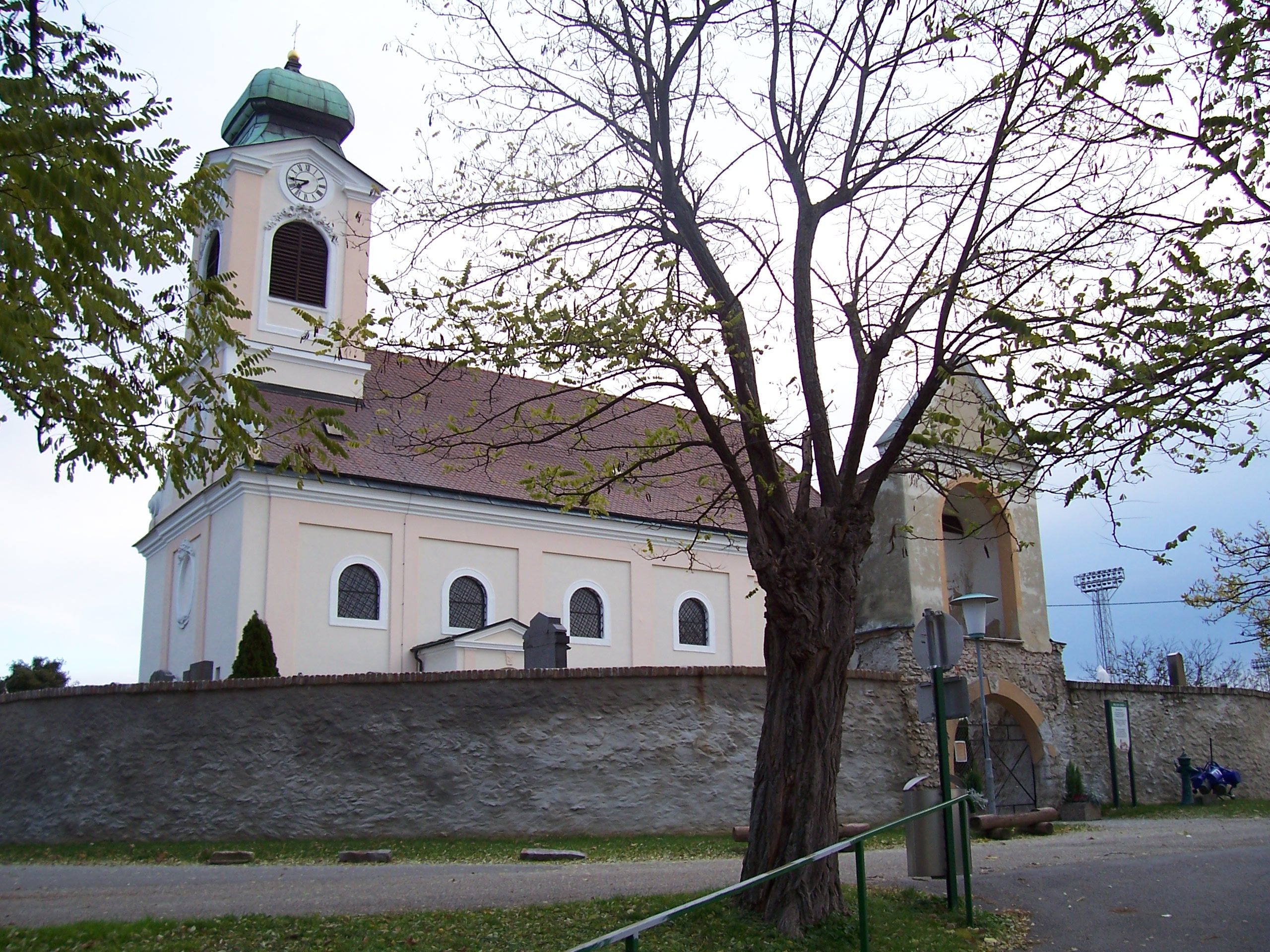 Kirche mit Turm und Uhr, umgeben von Bäumen und einer Steinmauer.
