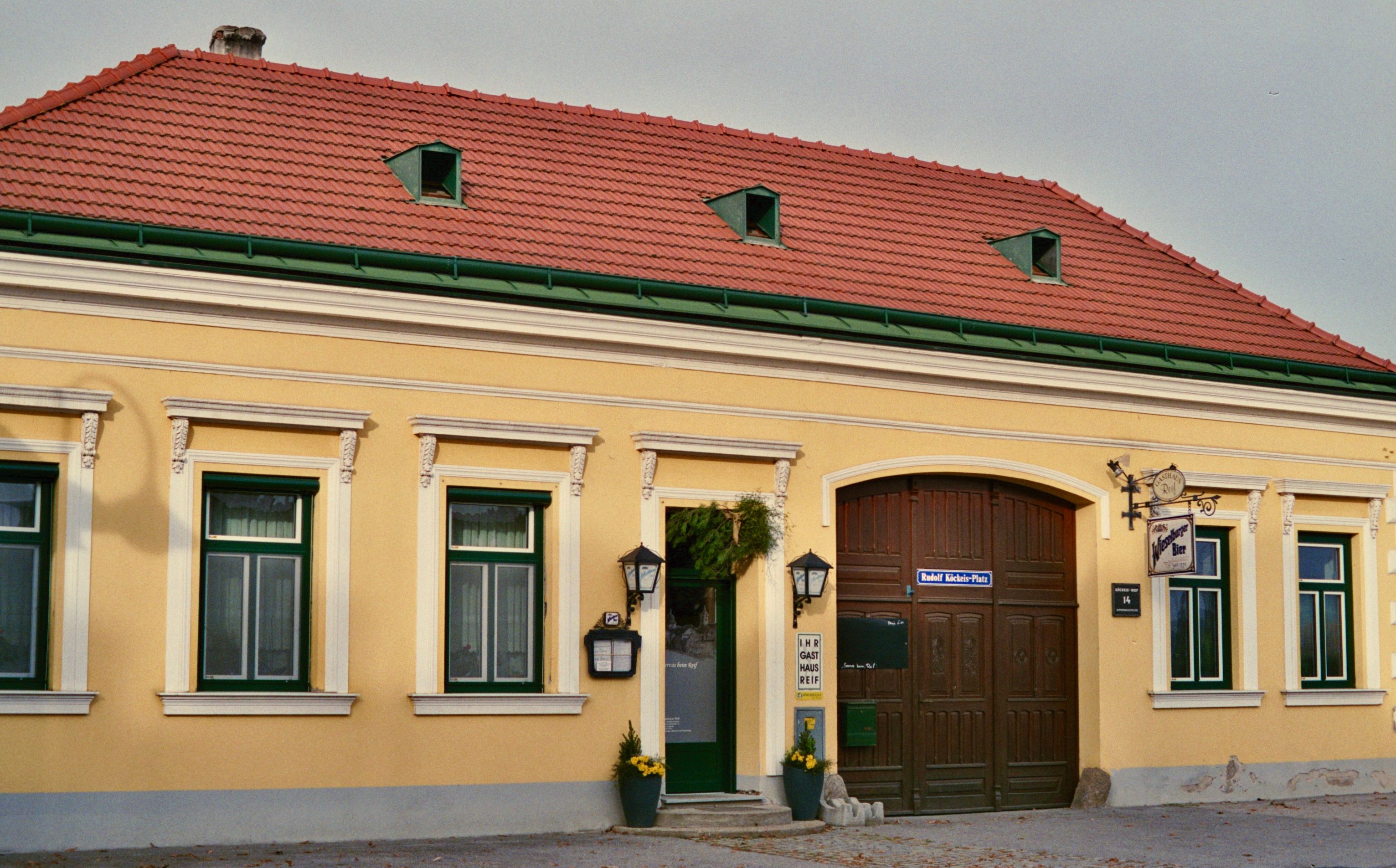 Außenansicht eines gelben Gasthauses mit roten Dachziegeln und grünen Fensterrahmen.