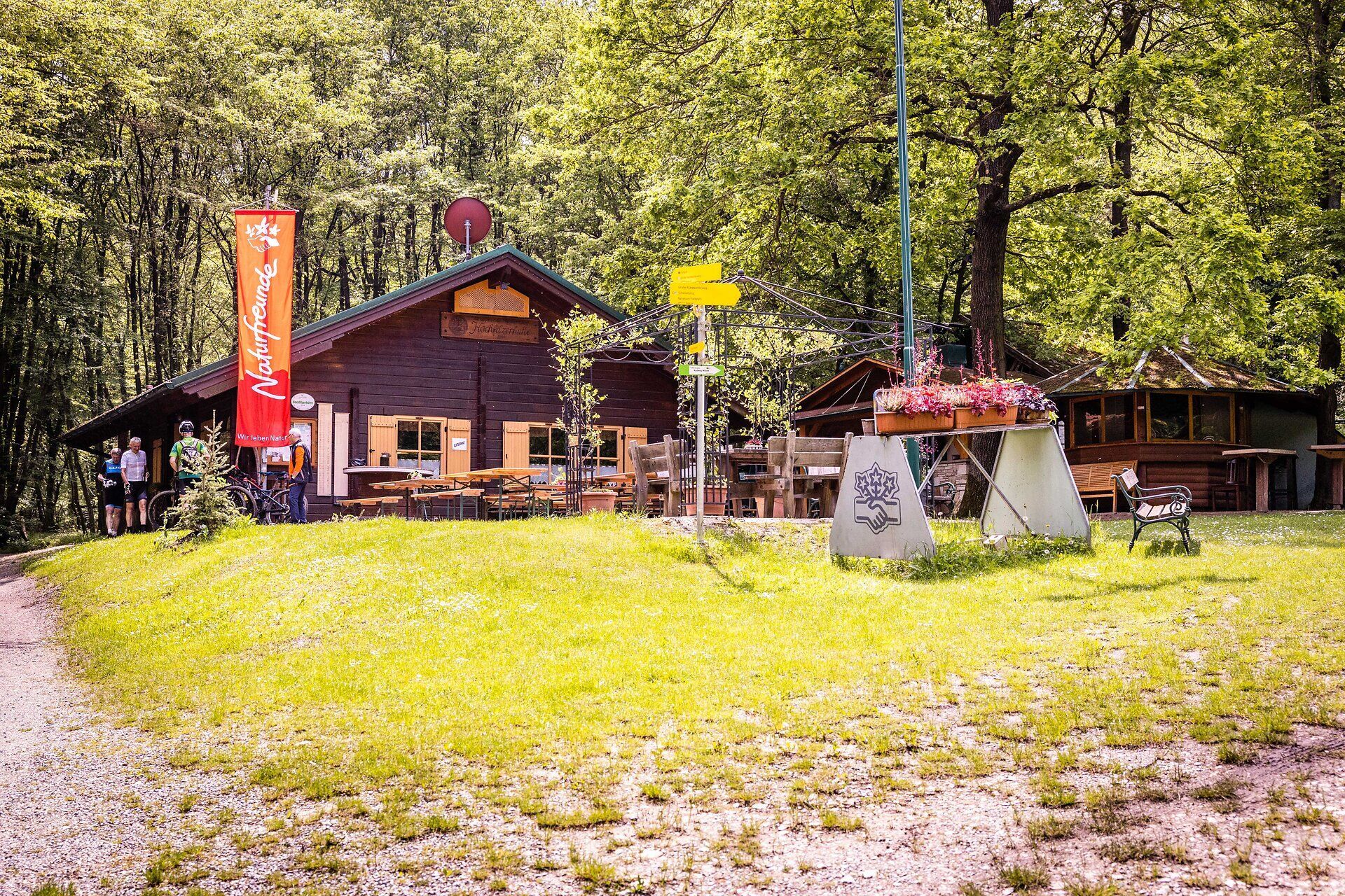Holzhütte der Naturfreunde mit Tischen, Bänken und Personen davor. 