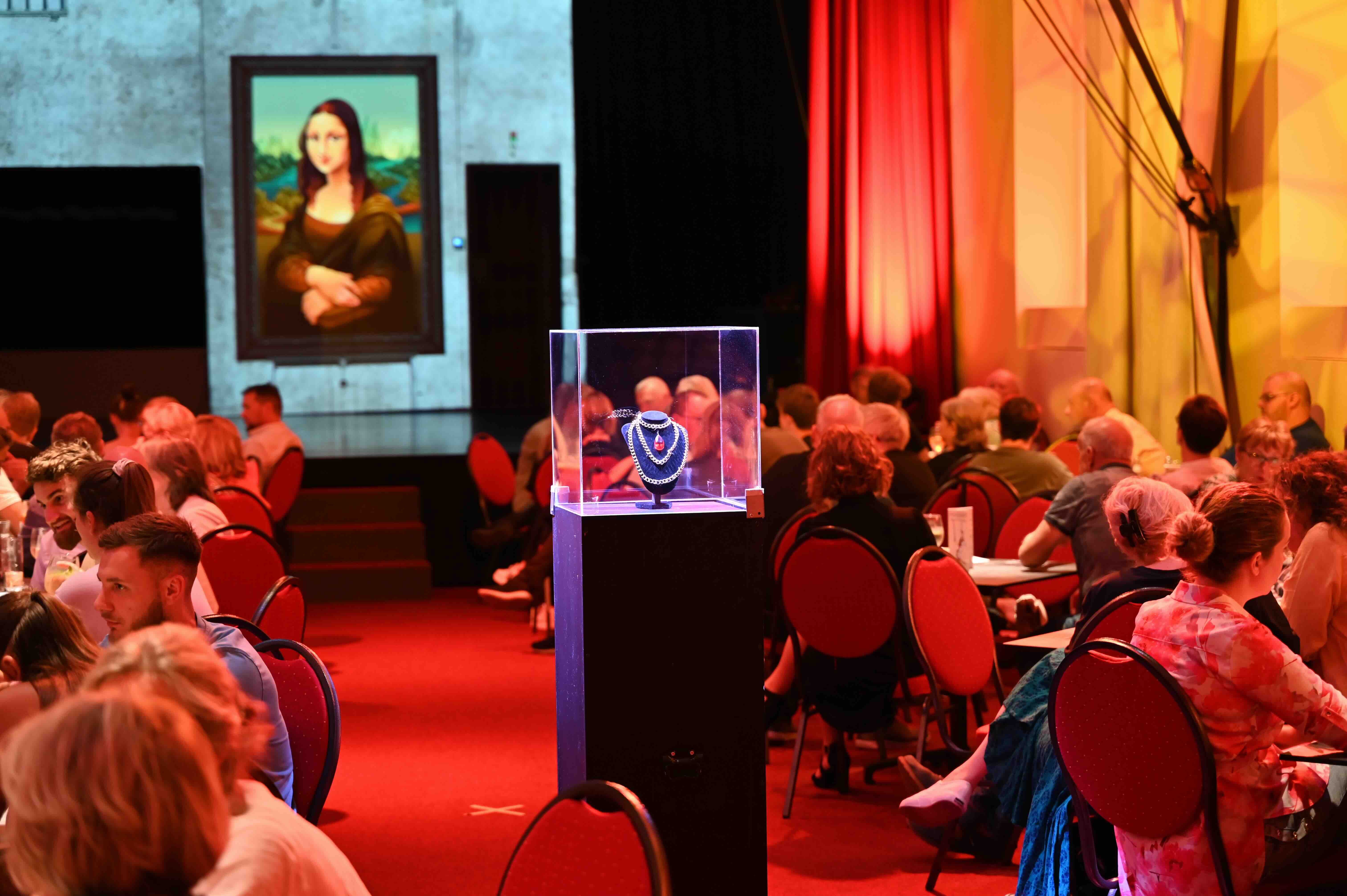 Ein Theater mit Menschen an Tischen, ein beleuchtetes Schmuckstück in einer Vitrine und ein Bild der Mona Lisa im Hintergrund.