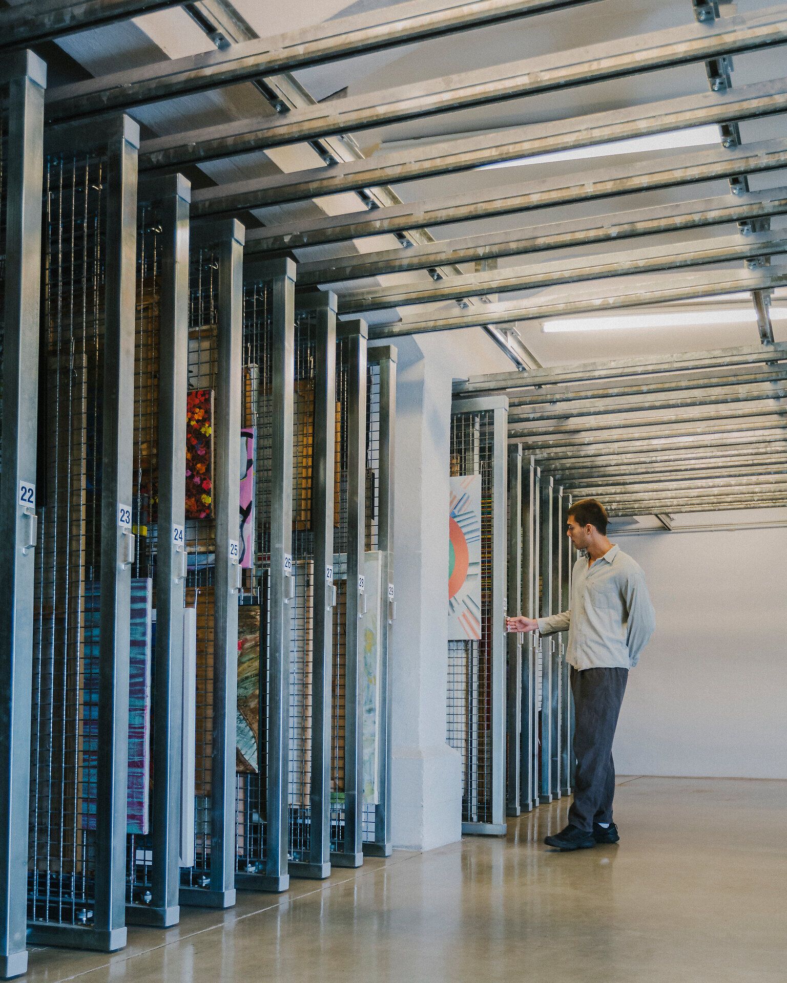Person steht in einem hellen Raum vor ausziehbaren, hohen Metallrahmen mit gelagerten Kunstwerken und betrachtet ein Bild.