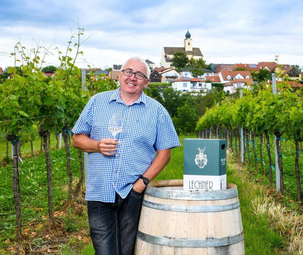 Man in a plaid shirt with a wine glass in a vineyard, in the background a village with a church.