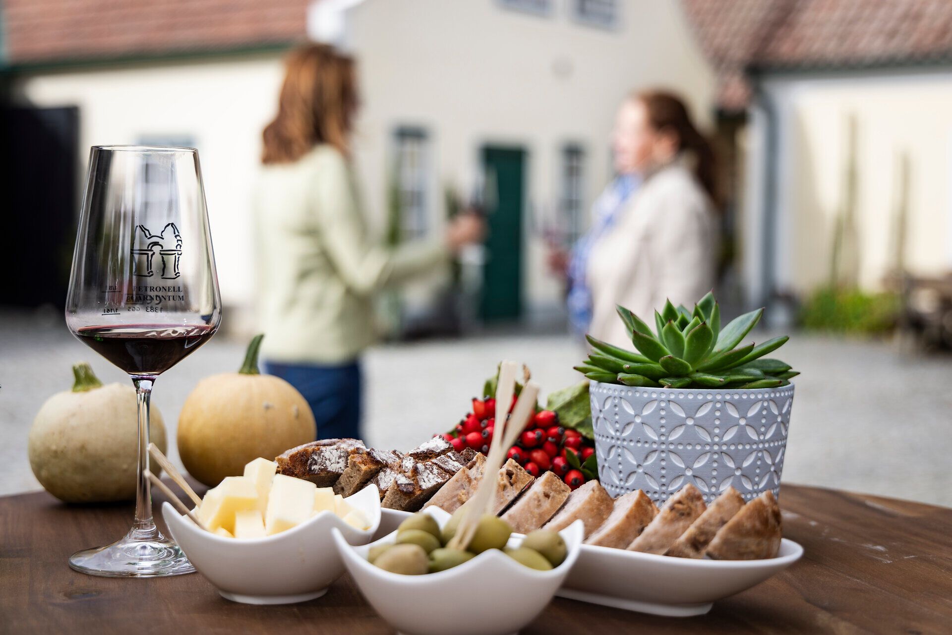 Rotweinglas, Brot, Oliven und Käsewürfel auf einem Holztisch. Im Hintergrund zwei Frauen mit Weingläsern. 