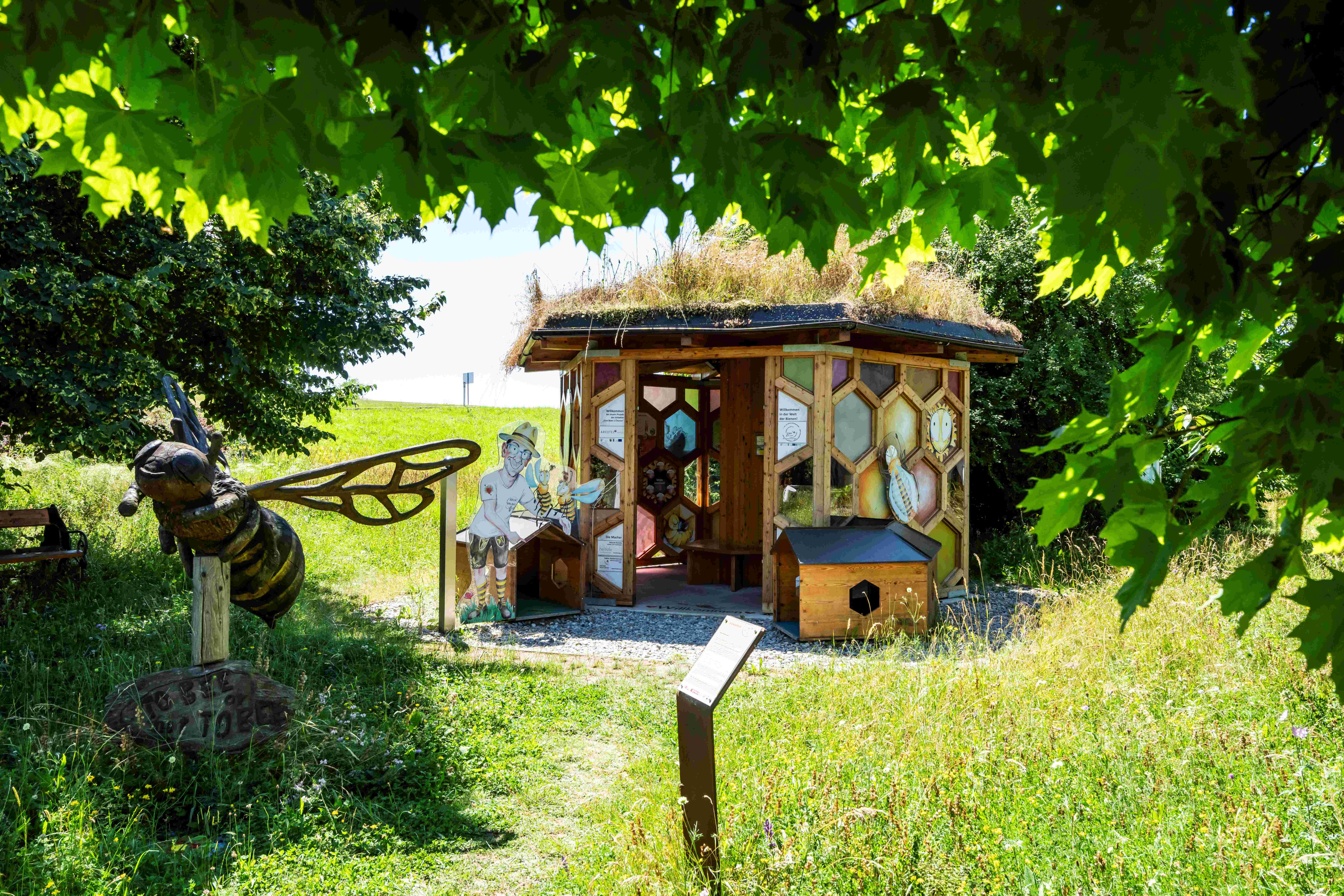 Ein Bienenpavillon mit Grasdach und Bienenfiguren im Bienenpark Klein-Pöchlarn, umgeben von grüner Natur.