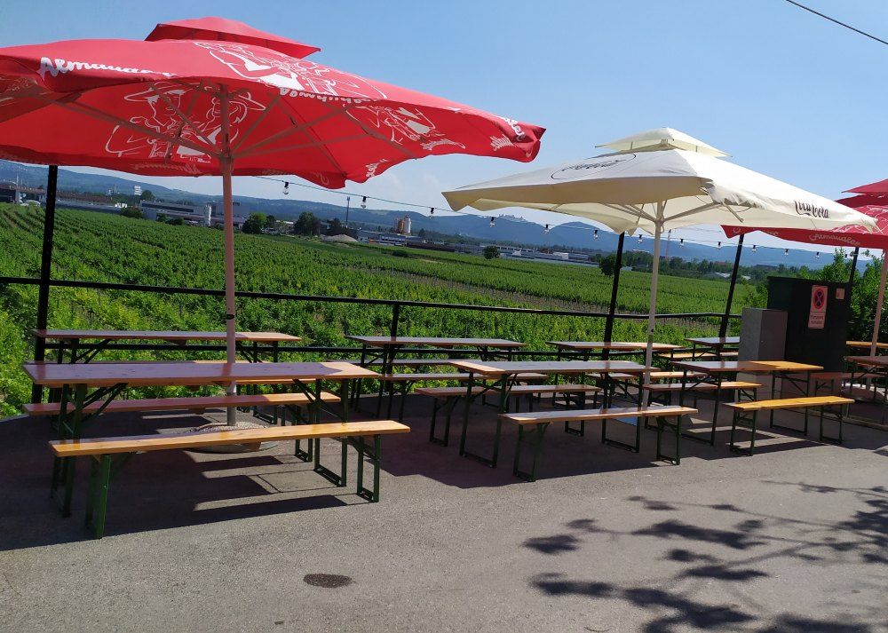 Biergarten mit Tischen und Sonnenschirmen vor einem Weinberg.