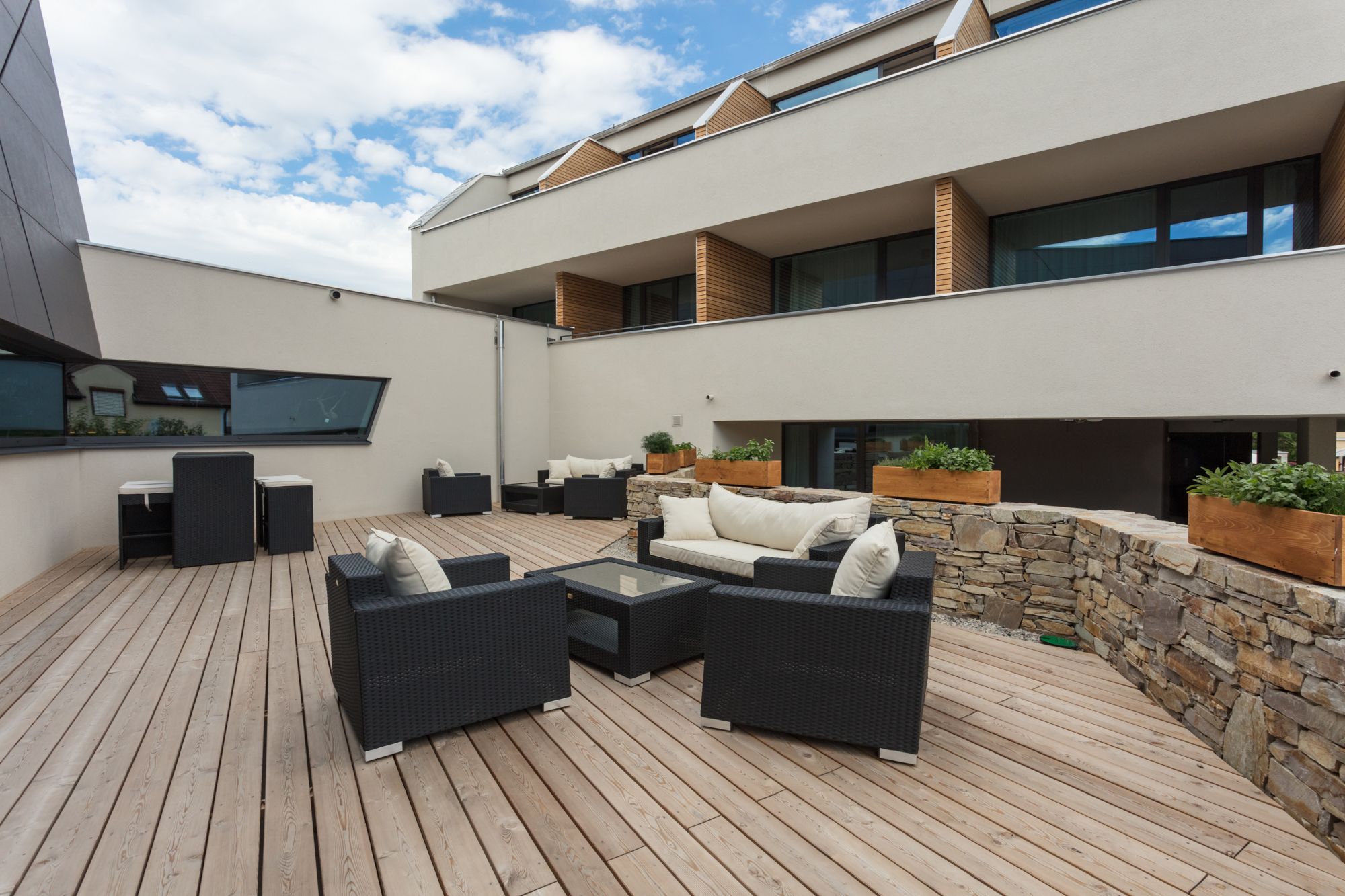 Moderne Terrasse mit Holzboden, schwarzen Rattanmöbeln und Steinmauer, umgeben von einem modernen Gebäude.