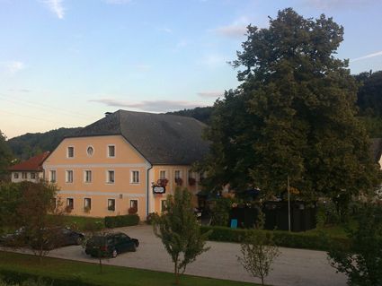 Ansicht eines gelben Gasthofs mit großem Baum und Parkplatz.