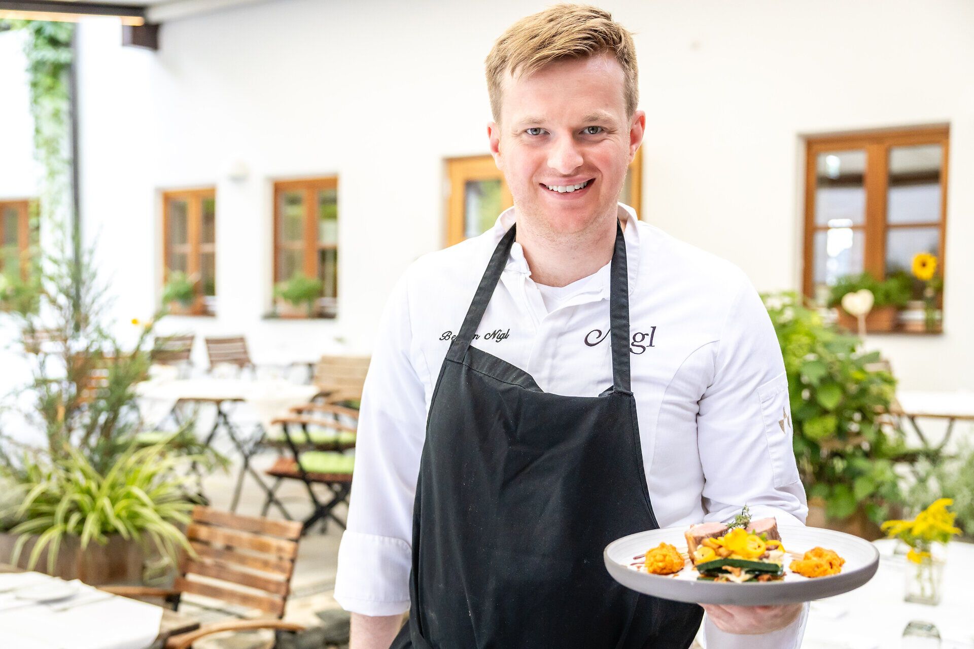 Herr Nigl hält ein weißes Teller mit einer Speise in der Hand und blickt in die Kamera.