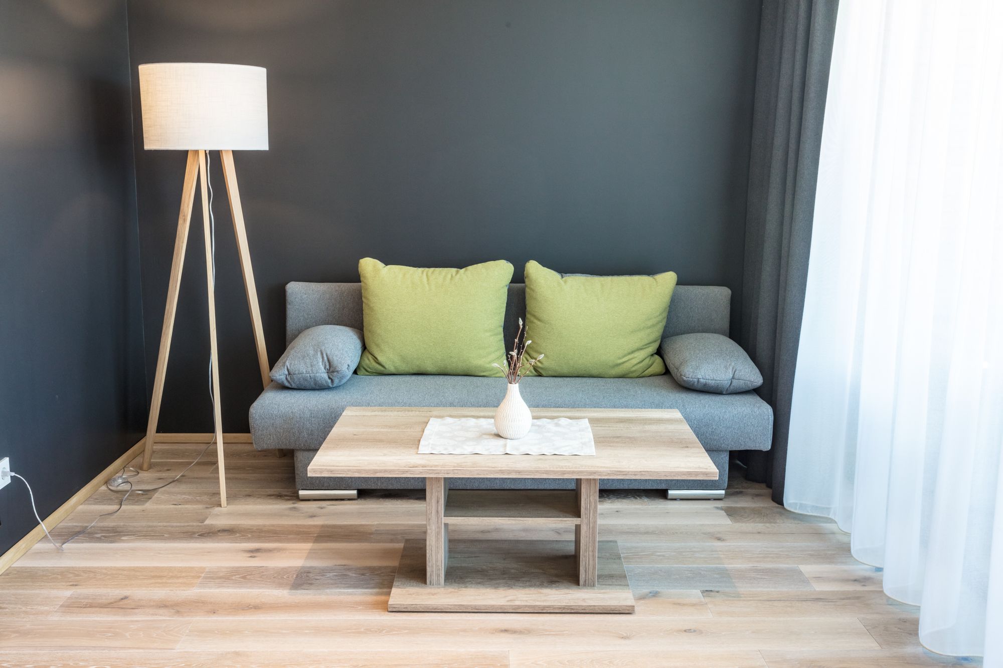 Modernes Wohnzimmer mit grauem Sofa, grünen Kissen, Holzlampe und Couchtisch vor dunkler Wand.