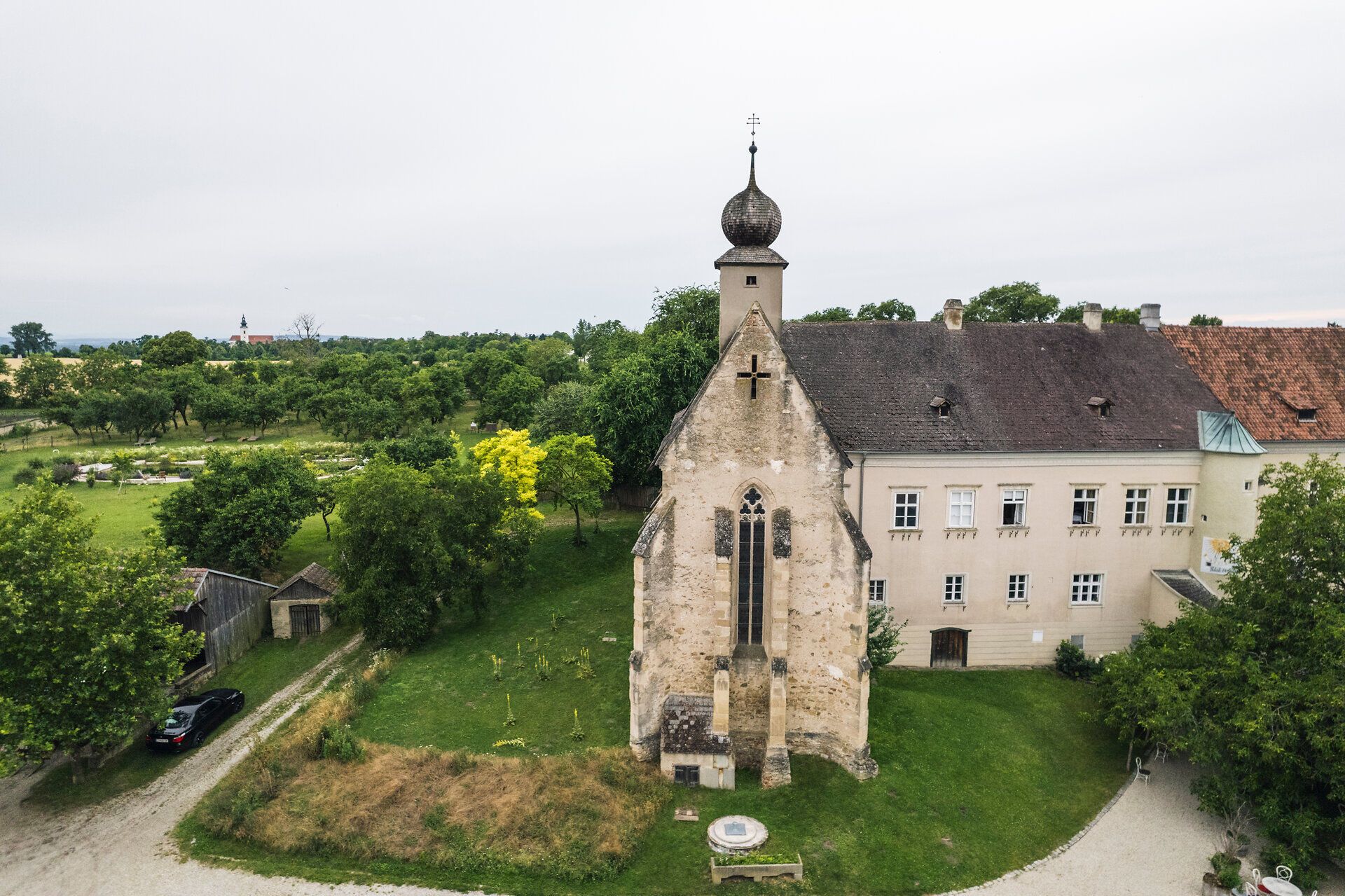 Alte Kirche mit Zweibelform-Spitze im Gut Oberstockstall mit einem klosterähnlichen Zubau, viel Grünfläche und Bäumen.