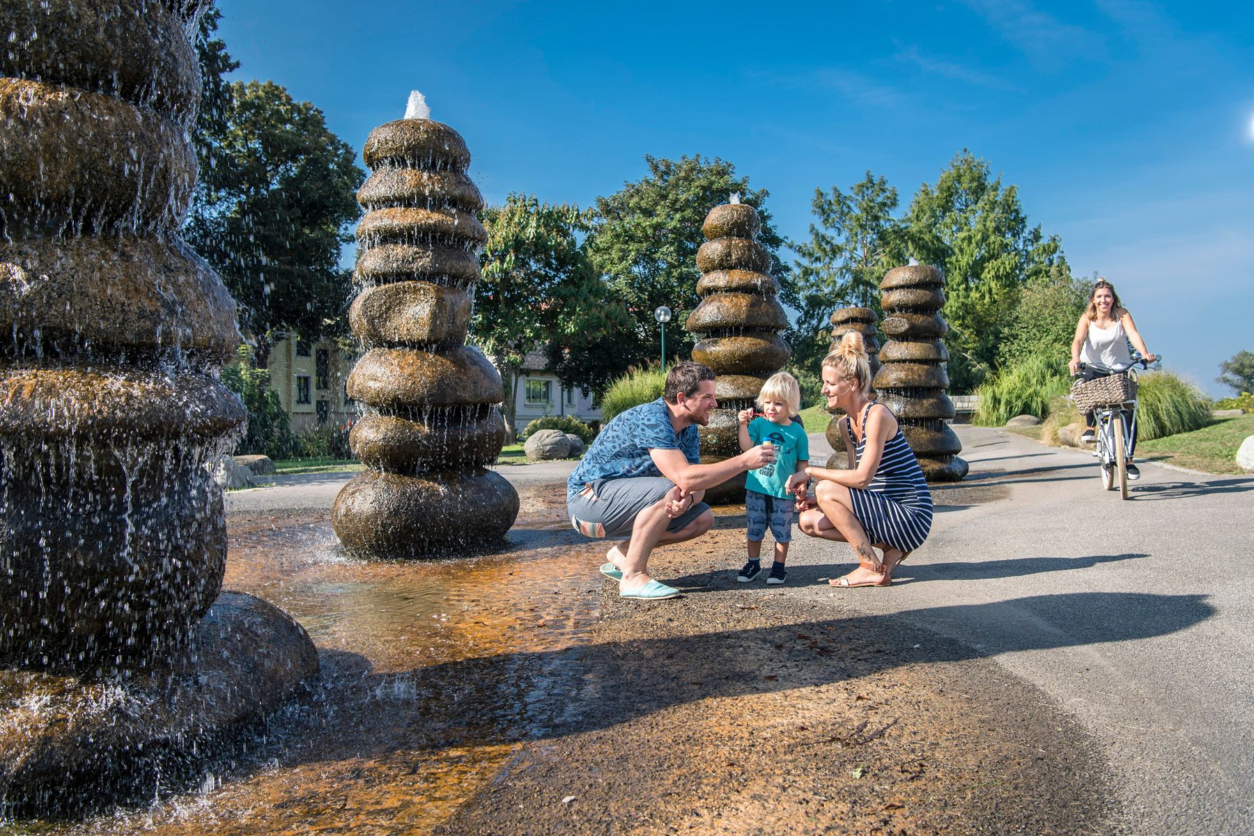 Familie am Brunnen in einem Park, mit einer Frau auf einem Fahrrad im Hintergrund.