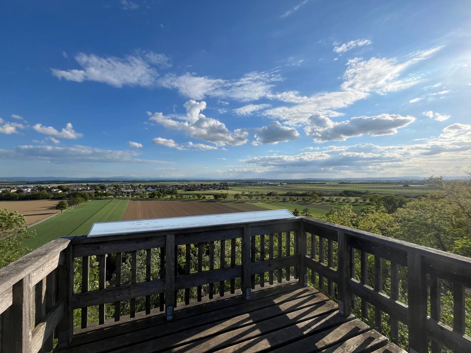 Holzaussichtsplattform mit Geländer über einer weiten Landschaft aus Feldern und kleinen Ortschaften. Darüber spannt sich ein großer blauer Himmel mit zahlreichen weißen Wolken, die von der tief stehenden Sonne beleuchtet werden. Die Szene vermittelt Weite und Ruhe.
