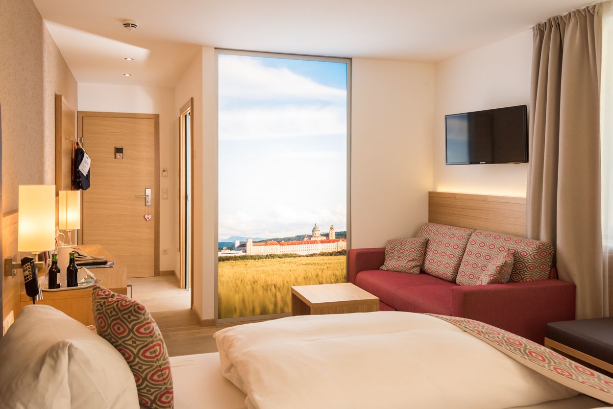 Modernes Hotelzimmer mit Doppelbett, rotem Sofa, Flachbildfernseher und großem Fenster mit Landschaftsausblick.