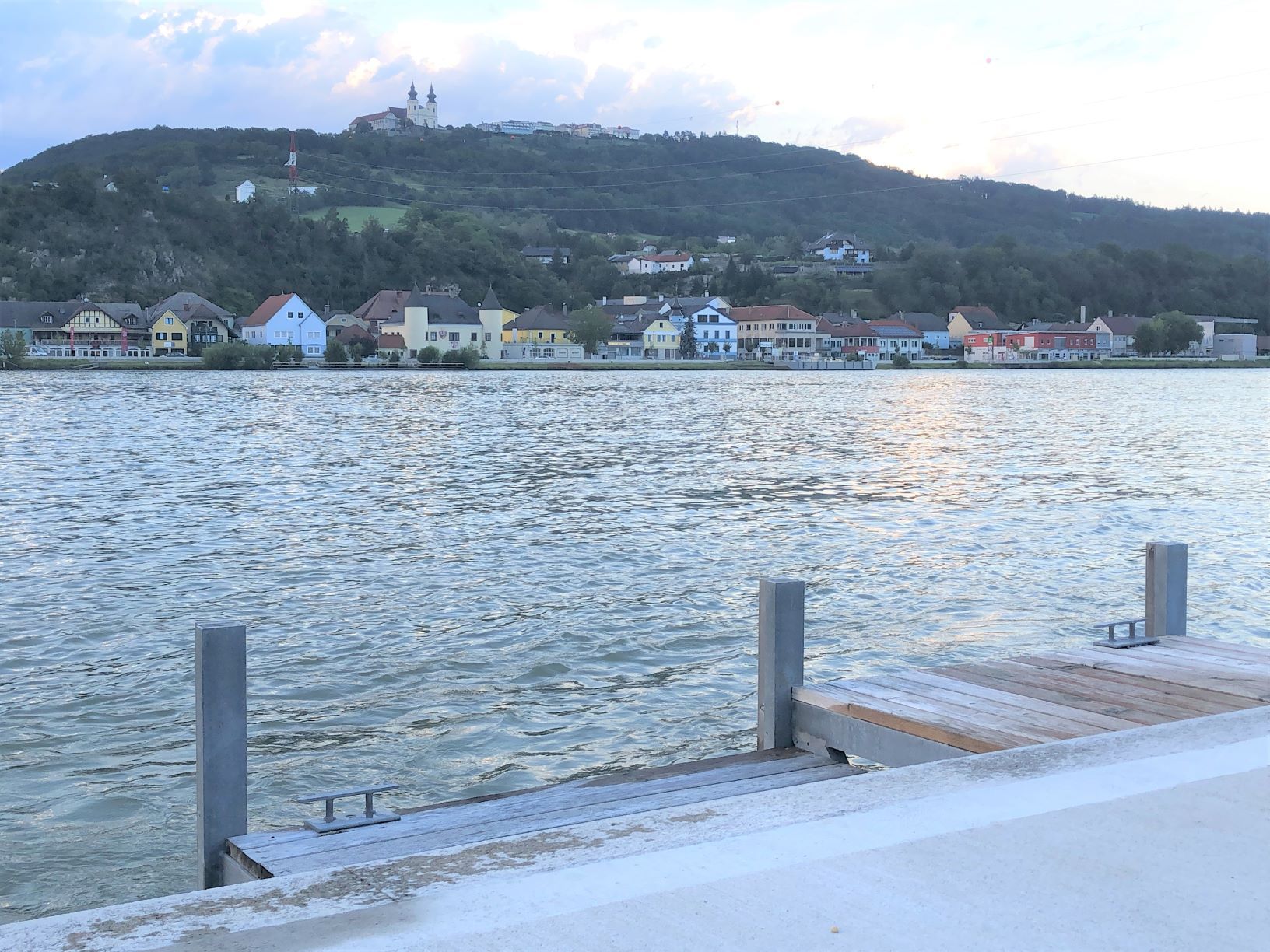 Blick von einer Anlegestelle auf den Fluss und bunte Häuser am gegenüberliegenden Ufer, mit einer Kirche auf einem Hügel im Hintergrund.