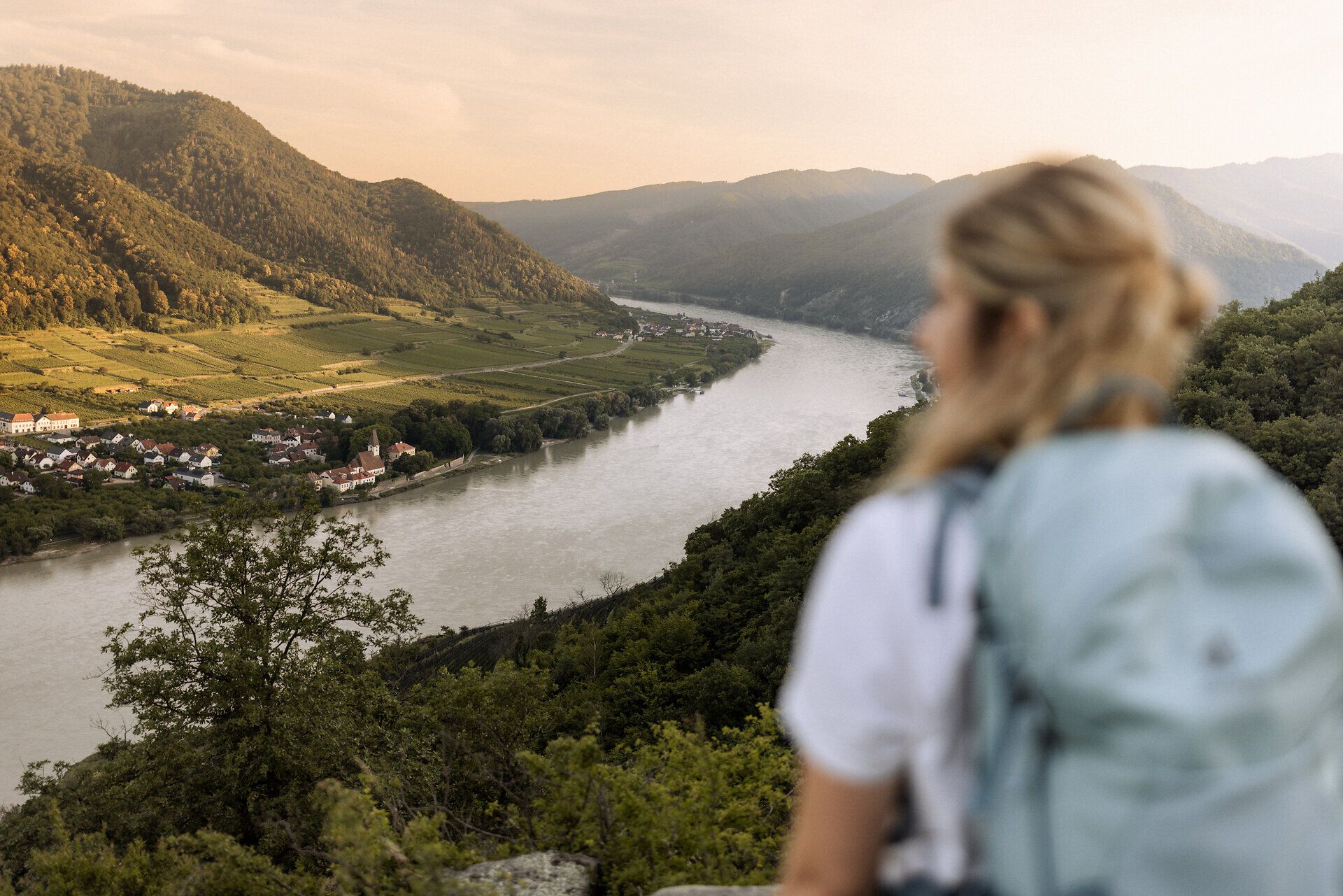 Eine Frau mit Wanderrucksack, die nur unscharf zu erkennen ist, steht auf einem Felsen und blickt auf das Donautal auf Höhe von Spitz an der Donau.