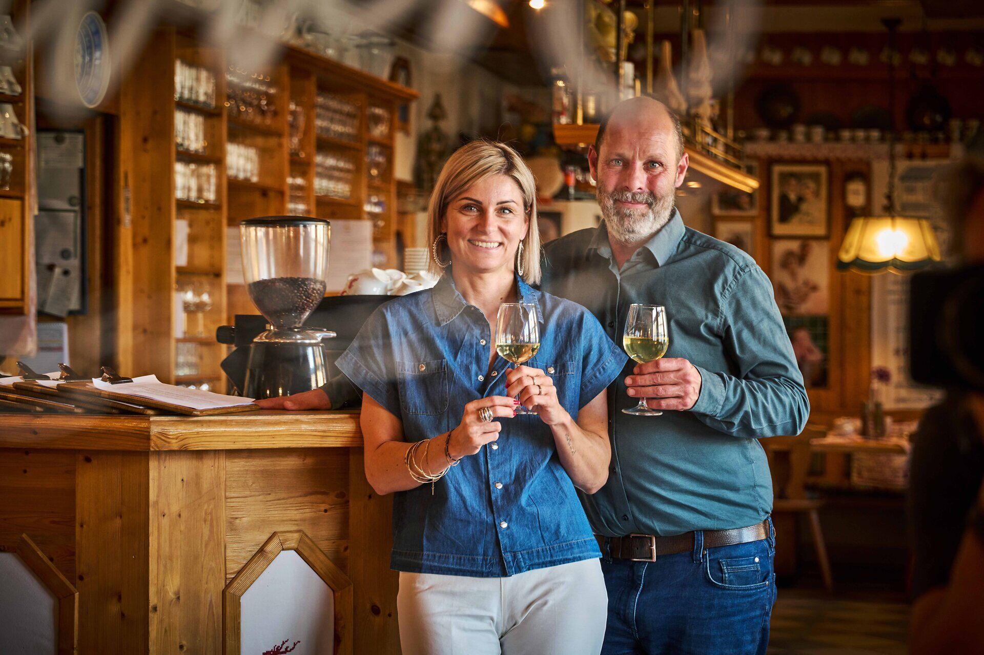 Eine lächelnde Frau und ein lächelnder Mann stehen mit je einem Glas Weißwein vor der Holztheke eines Gasthauses.