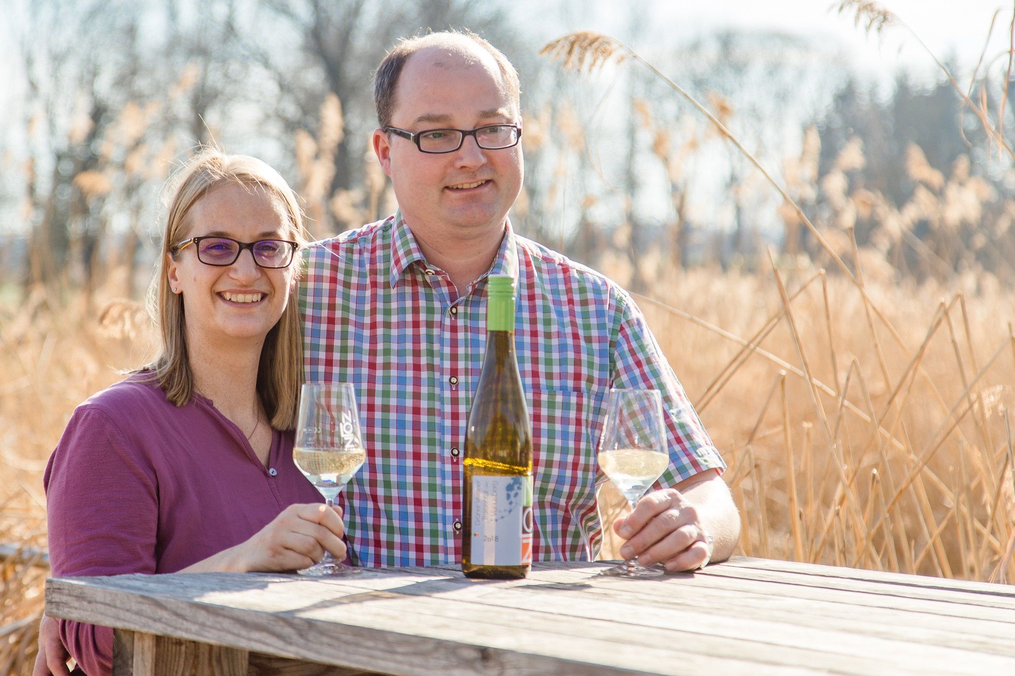 Ein Mann und eine Frau sitzen an einem Holztisch im Freien mit Weingläsern und einer Weinflasche.