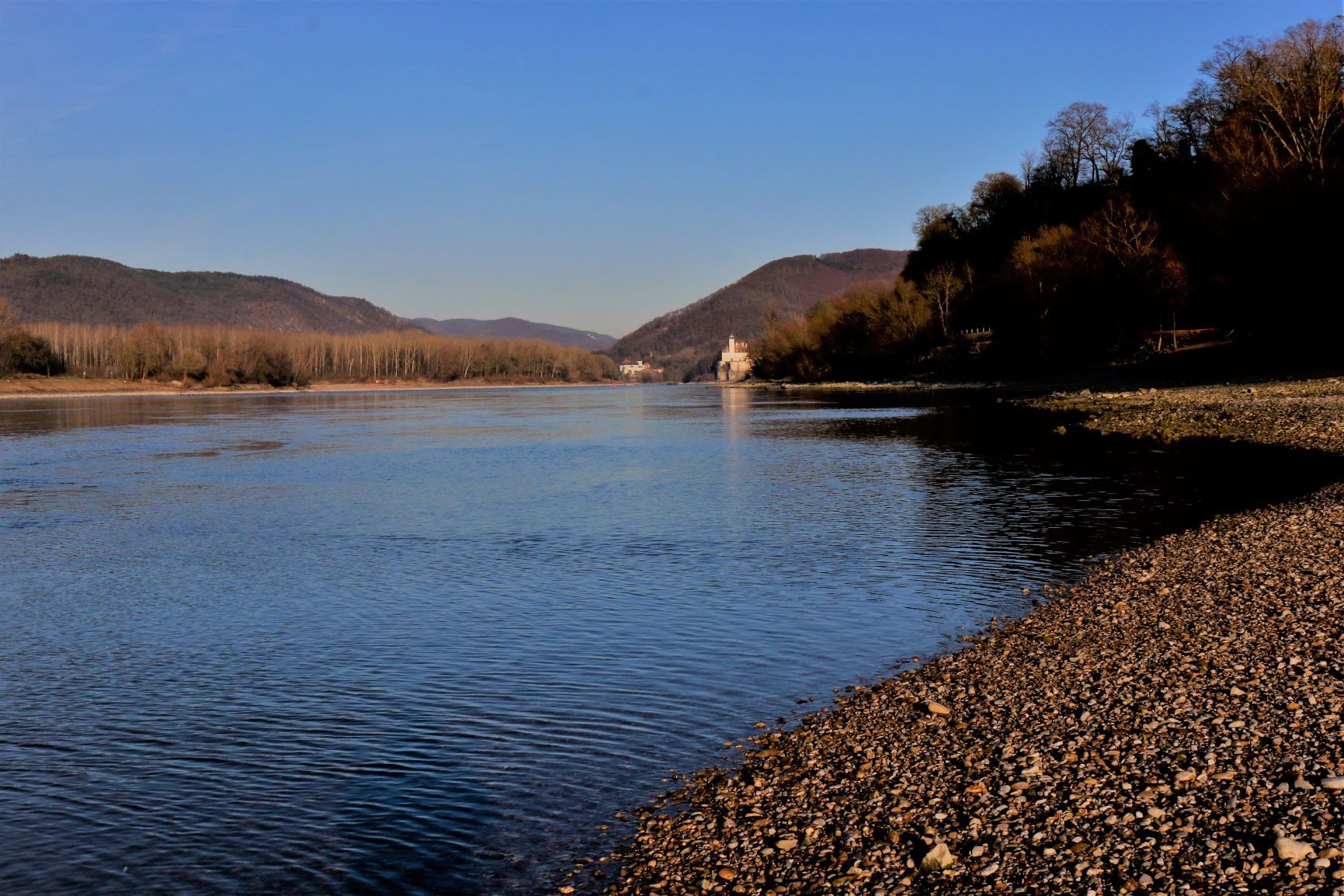 Flussufer mit Kiesstrand und bewaldeten Hügeln im Hintergrund.
