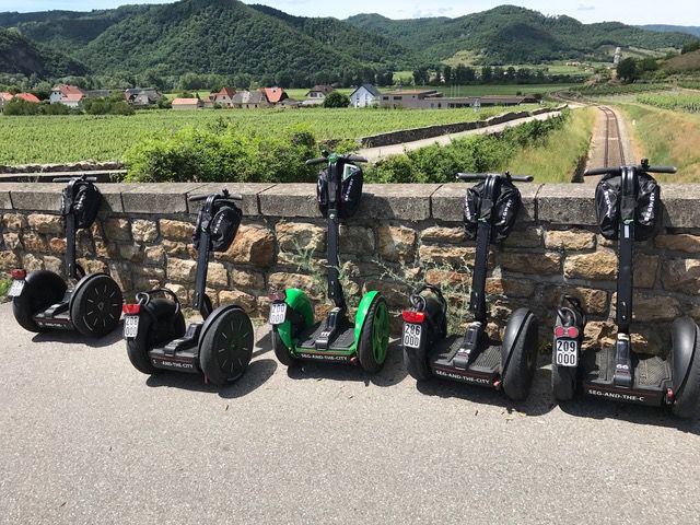 Six Segways stand in front of a stone wall with a landscape in the background.