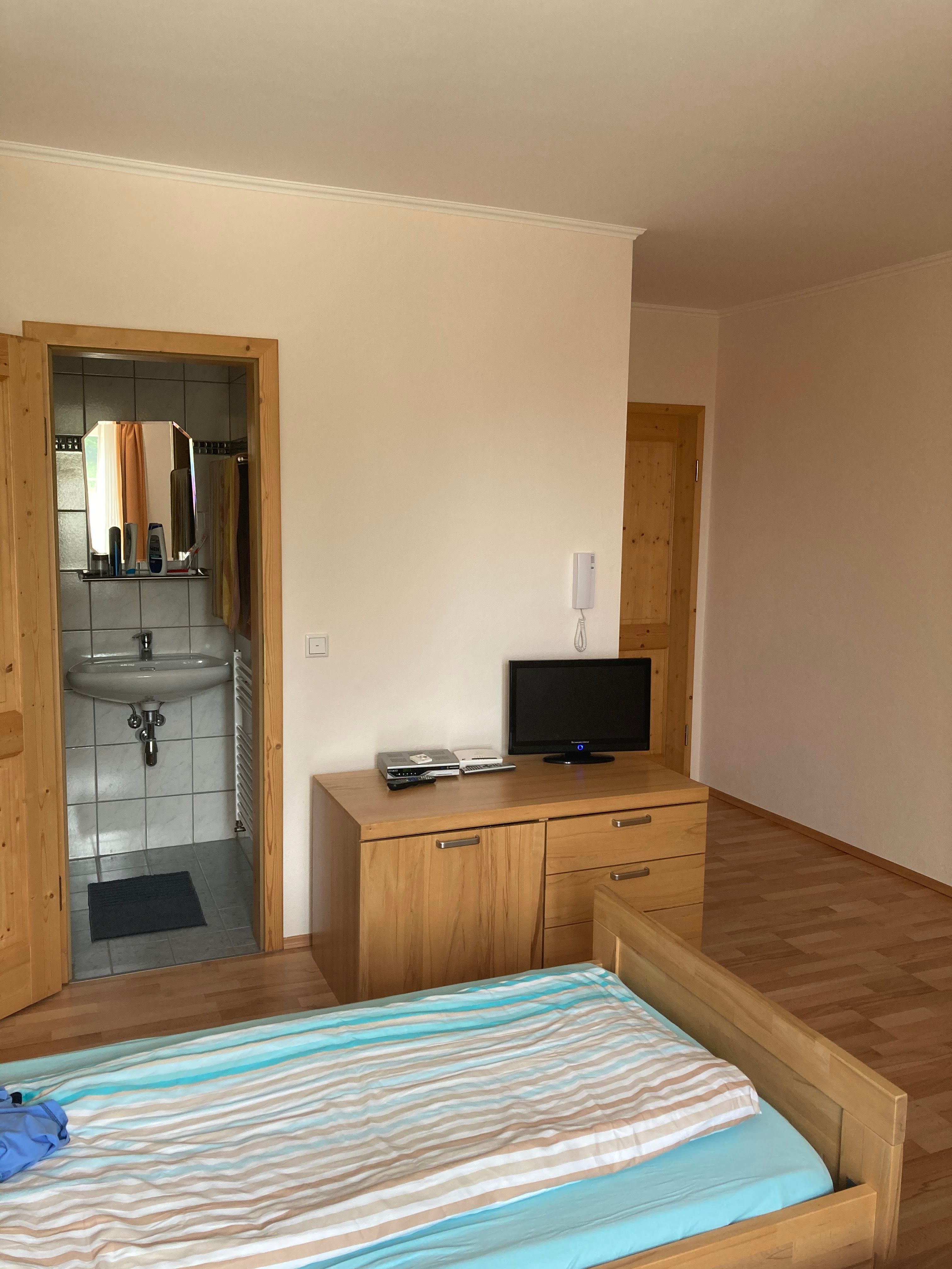 Zimmer mit einem Einzelbett, einem Sideboard mit Fernseher. Im Hintergrund die Türe zum Badezimmer.