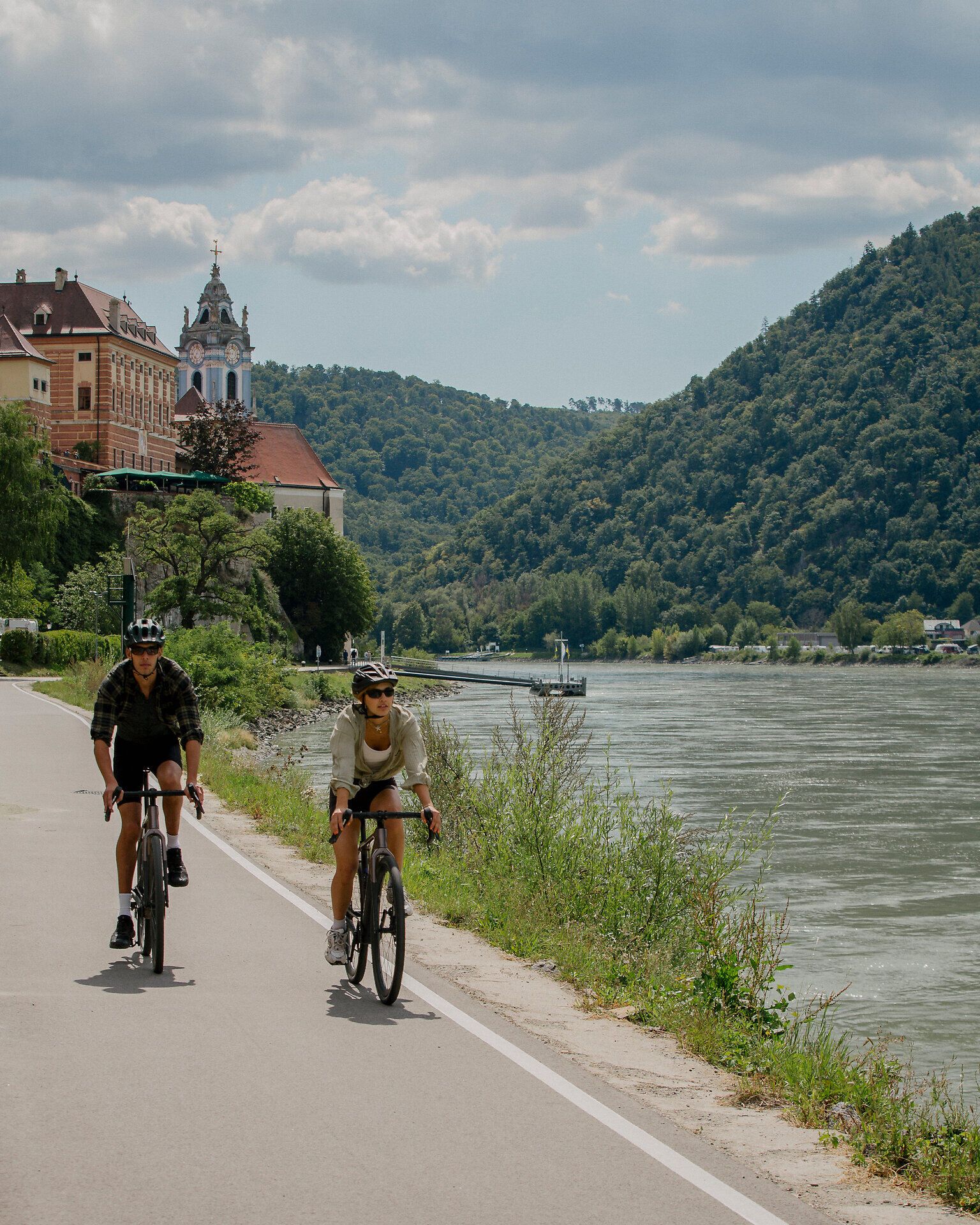 Zwei Radfahrende fahren nebeneinander auf einem gepflasterten Radweg entlang des Donauufers. Im Hintergrund ist der Blaue Turm des Stift Dürnsteins zu sehen, eingebettet in eine malerische Landschaft aus bewaldeten Hügeln und dem ruhig fließenden Fluss. Der Himmel ist leicht bewölkt und schafft eine entspannte Atmosphäre.