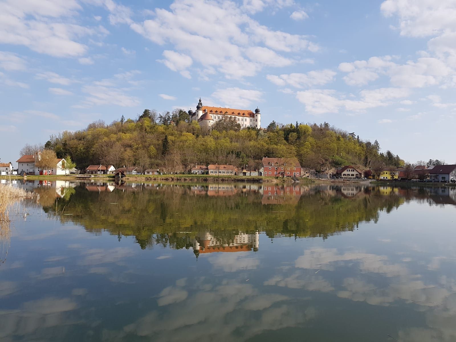 Ein Schloss auf einem Hügel spiegelt sich in einem ruhigen See, umgeben von Bäumen und kleinen Häusern.
