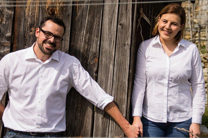Zwei Personen in weißen Hemden stehen vor einer Holzwand und lächeln.