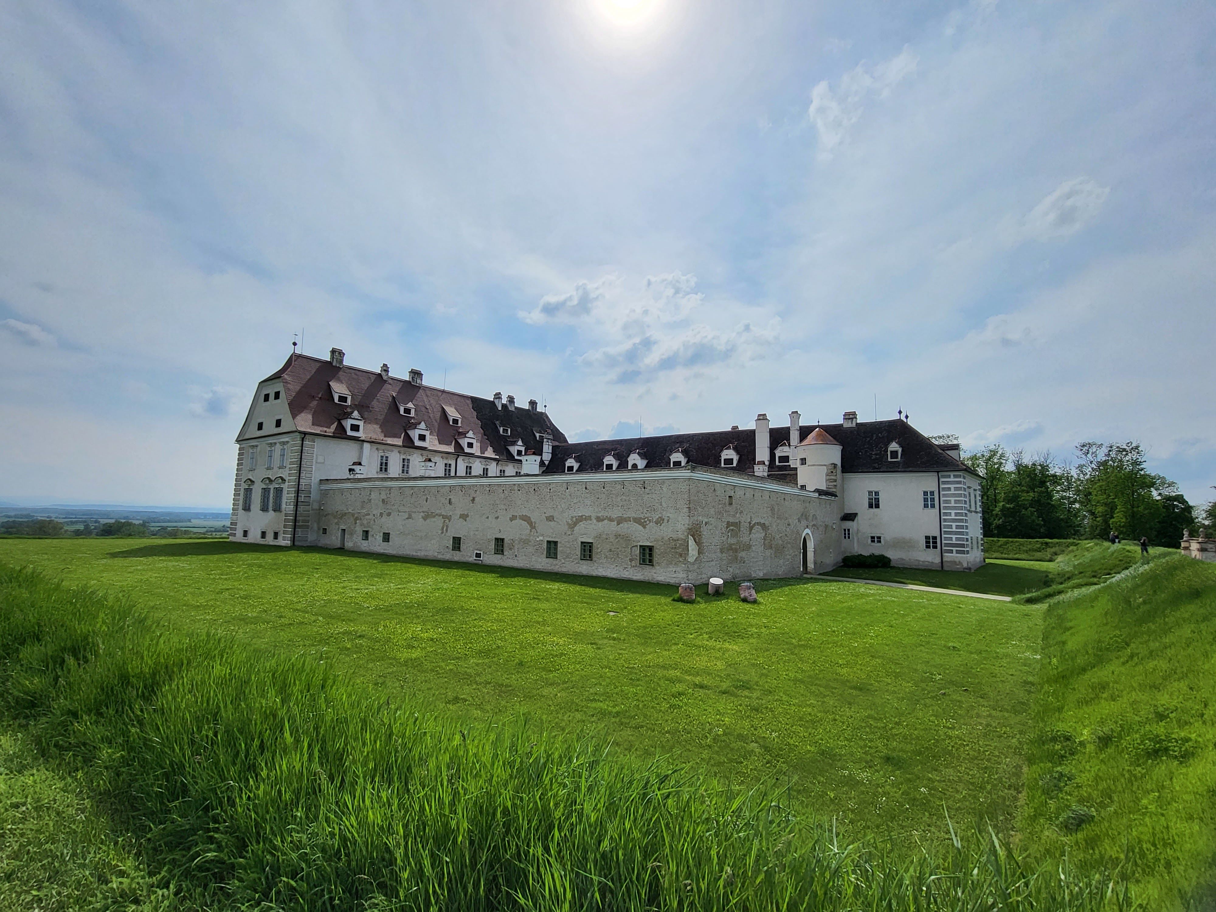 A large, historic castle with white walls and red roofs stands on a green meadow under a cloudy sky.