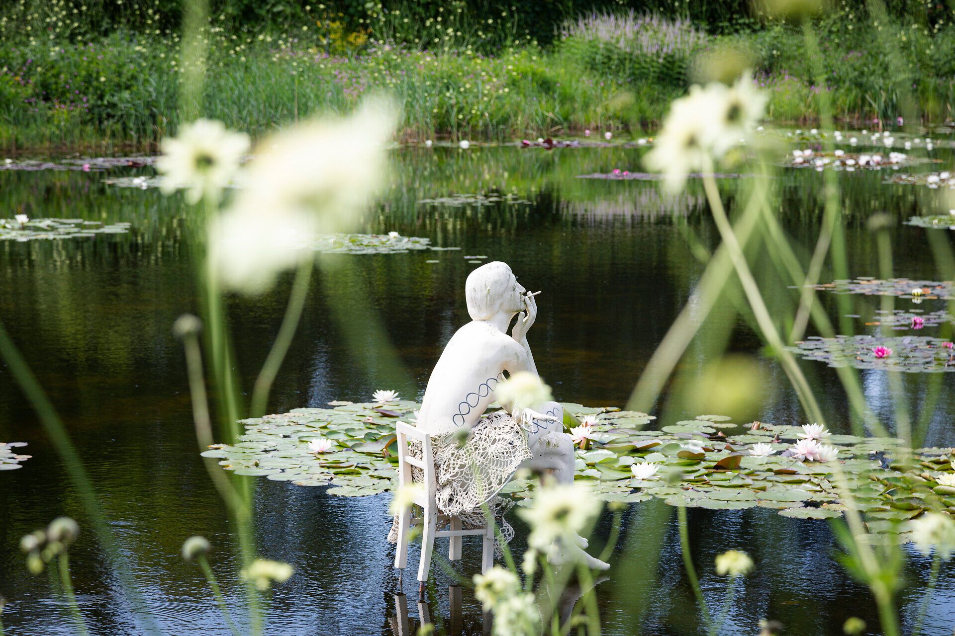 Inmitten der sanften Wellen des Wassers thront eine elegante Skulptur, die in stiller Kontemplation verweilt. Umgeben von blühenden Seerosen und dem sanften Rascheln der Gräser, lädt dieser Ort zum Verweilen und Träumen ein. Die harmonische Verbindung von Kunst und Natur schafft eine Oase der Ruhe und Inspiration.