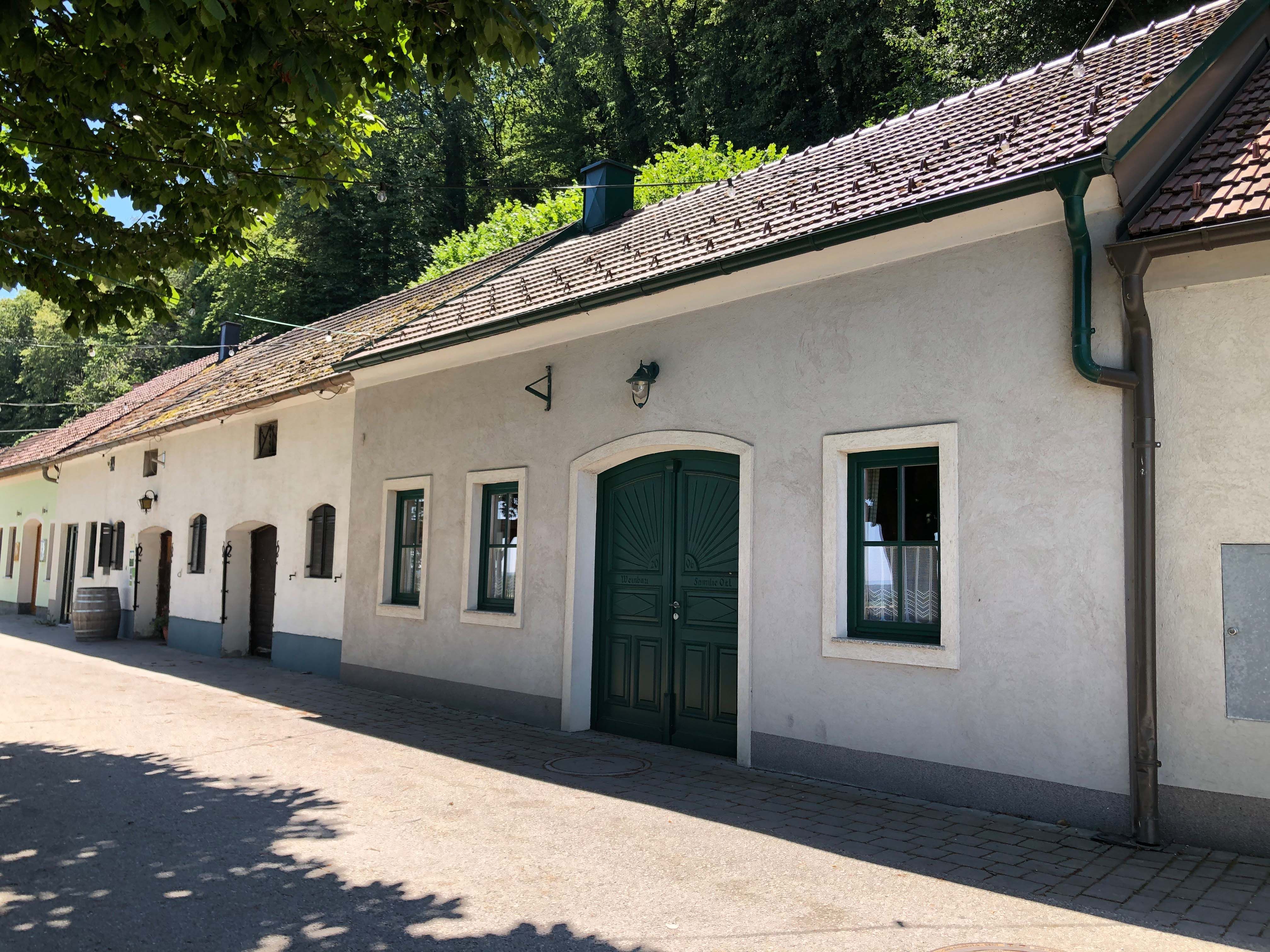 Außenansicht eines traditionellen Heurigen mit weißen Wänden und grünem Tor, umgeben von Bäumen.