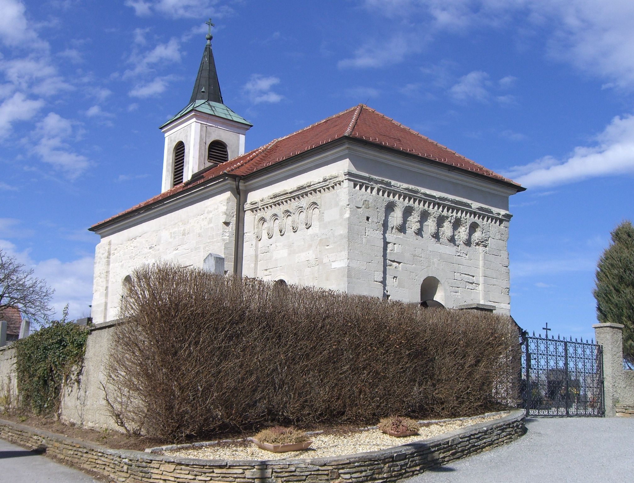 Romanische Kirche mit Glockenturm und roten Ziegeldach, umgeben von einer Mauer und Sträuchern.