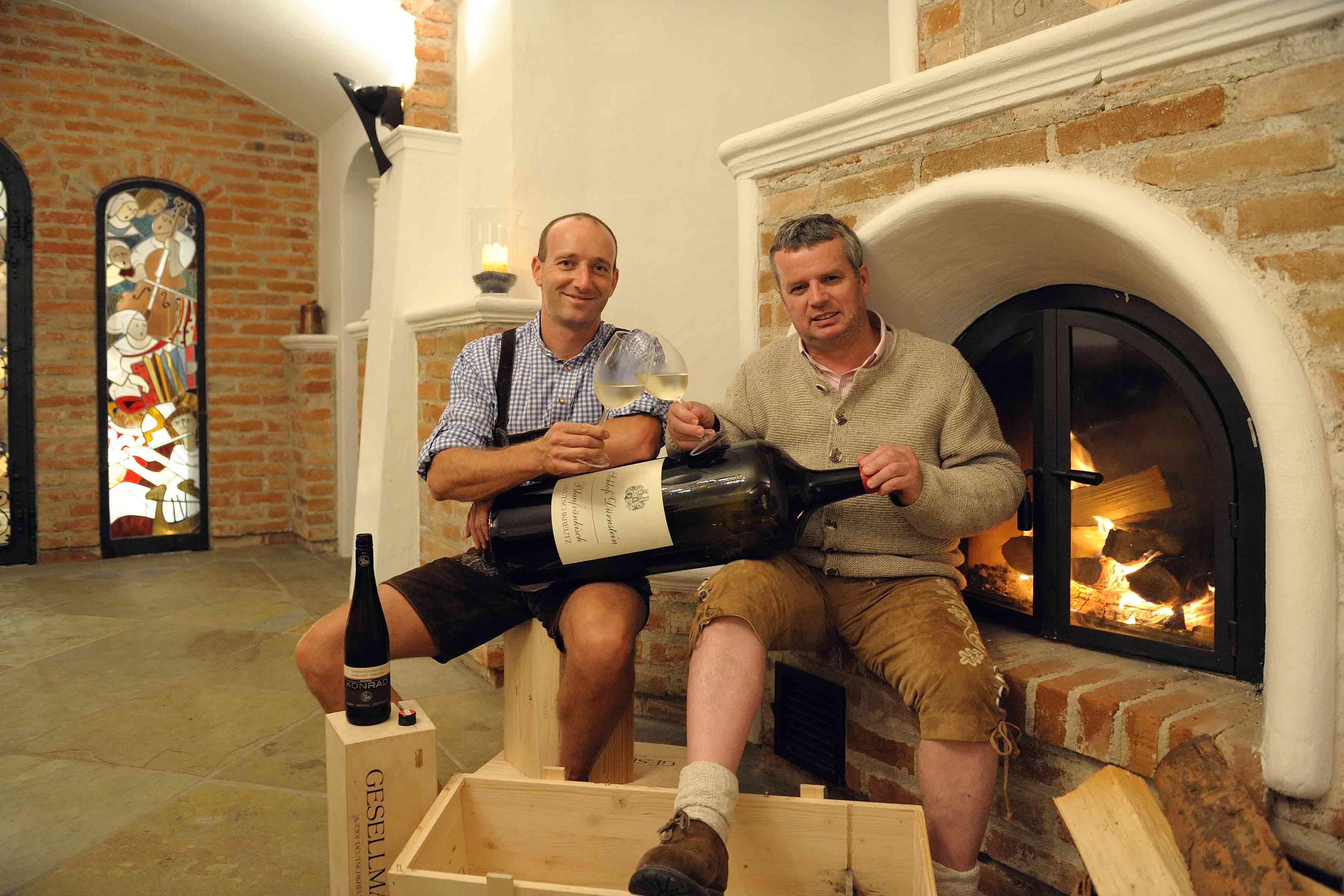 Zwei Männer in traditioneller Kleidung halten große Weinflasche vor Kamin.