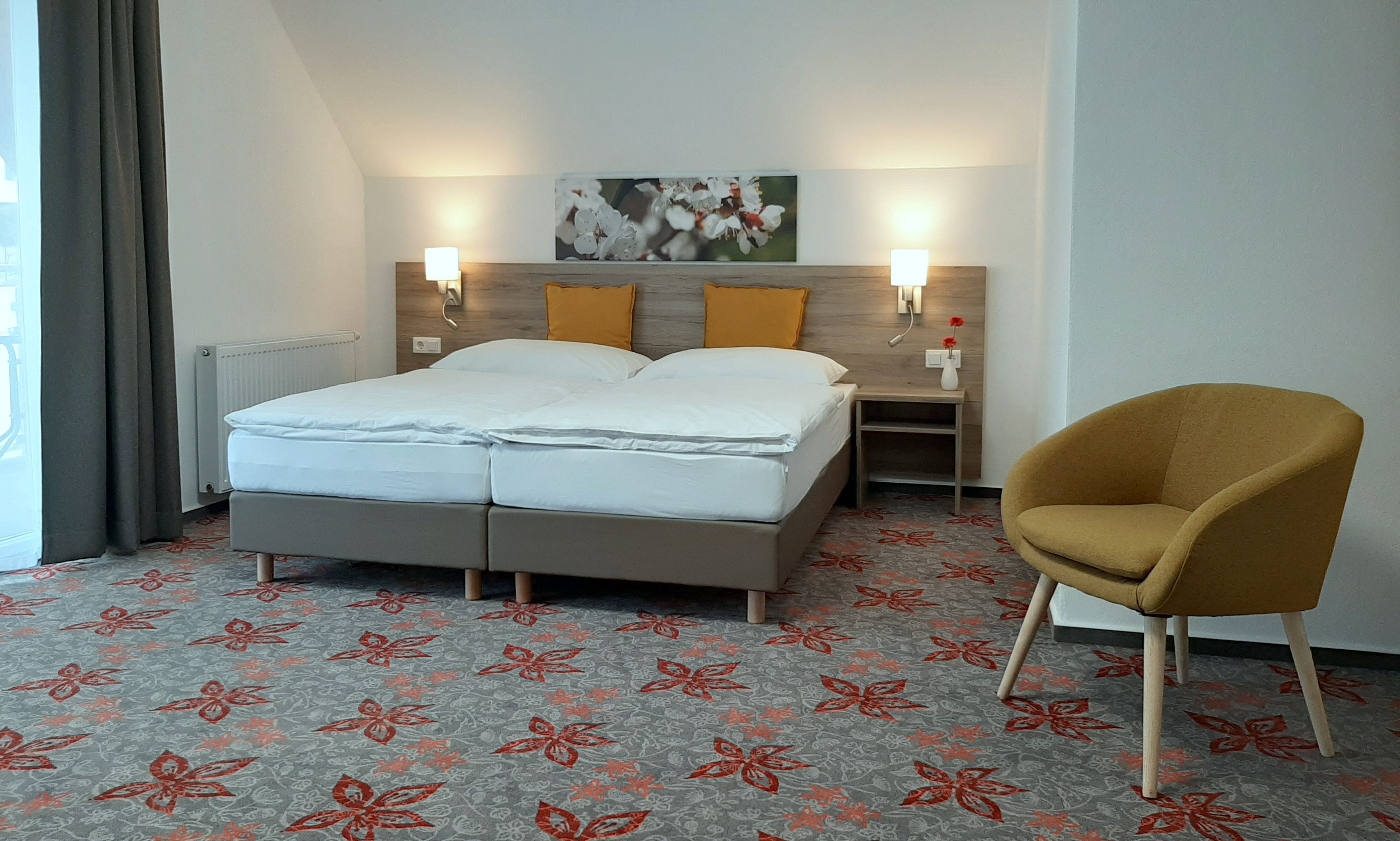 Modernes Doppelzimmer mit zwei Betten, gelben Kissen, Teppich mit Blumenmuster und einem gelben Sessel.