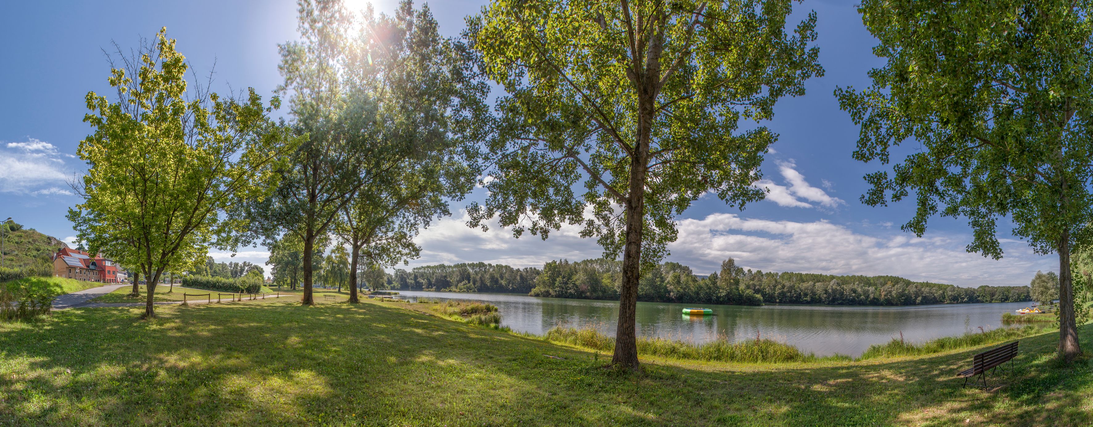Panoramablick auf einen See mit Bäumen und einer Bank im Vordergrund, sonniger Himmel.