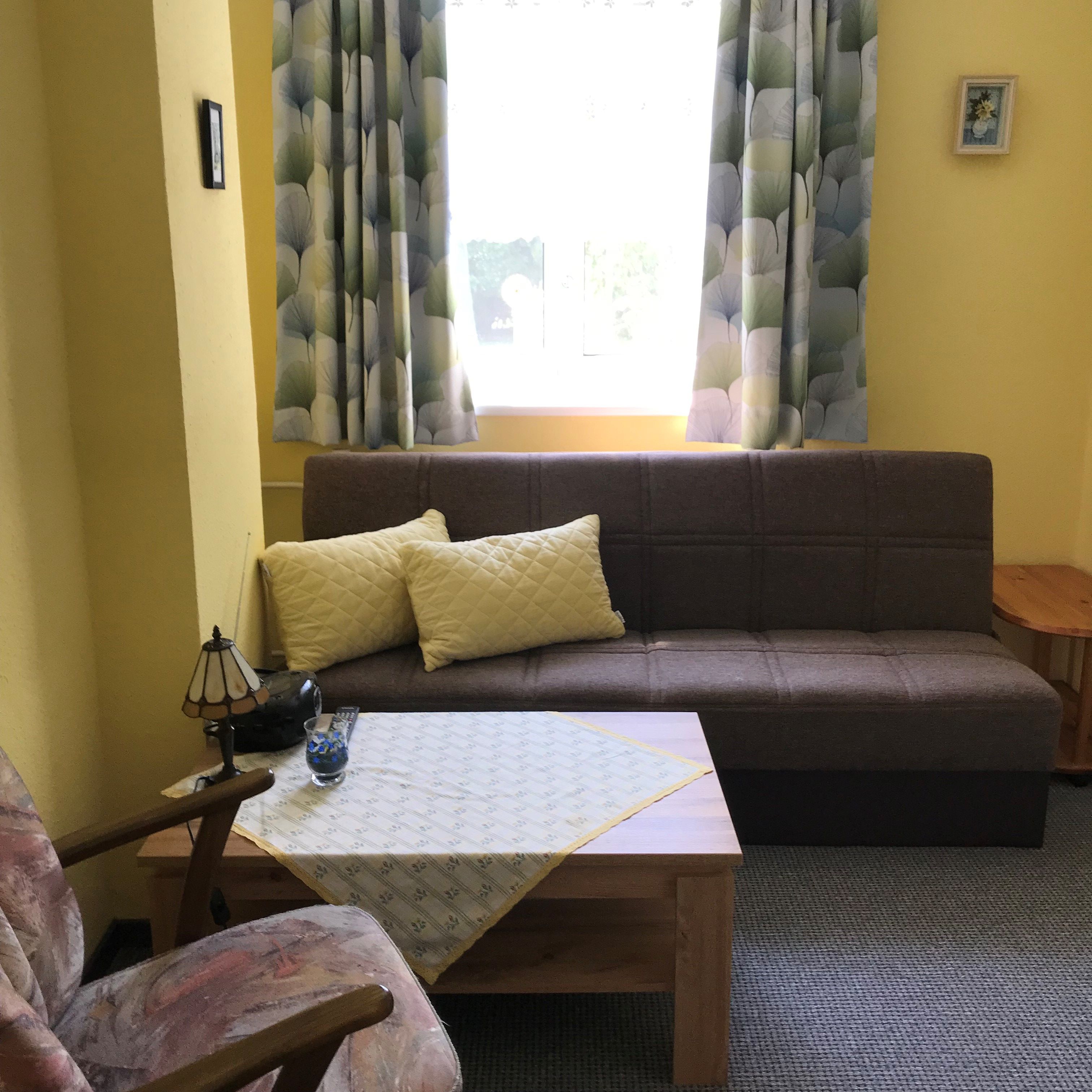 Wohnzimmer mit braunem Sofa, gelben Kissen, Tisch mit Tischdecke, Sessel und Fenster mit gemusterten Vorhängen.