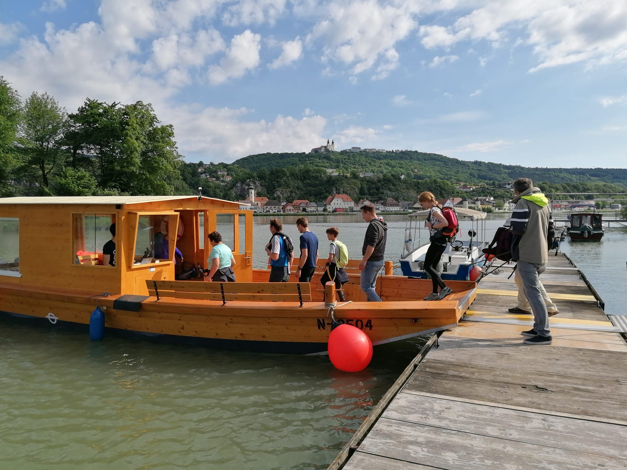 Menschen steigen auf ein Holzboot an einem Steg ein, im Hintergrund eine hügelige Landschaft mit Gebäuden.