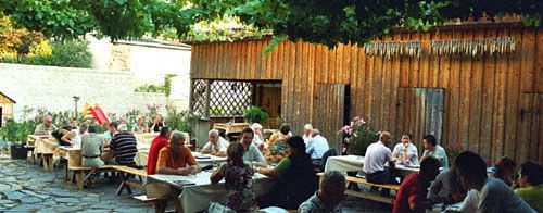 Menschen sitzen an Holztischen in einem gemütlichen Hof mit Weinreben.