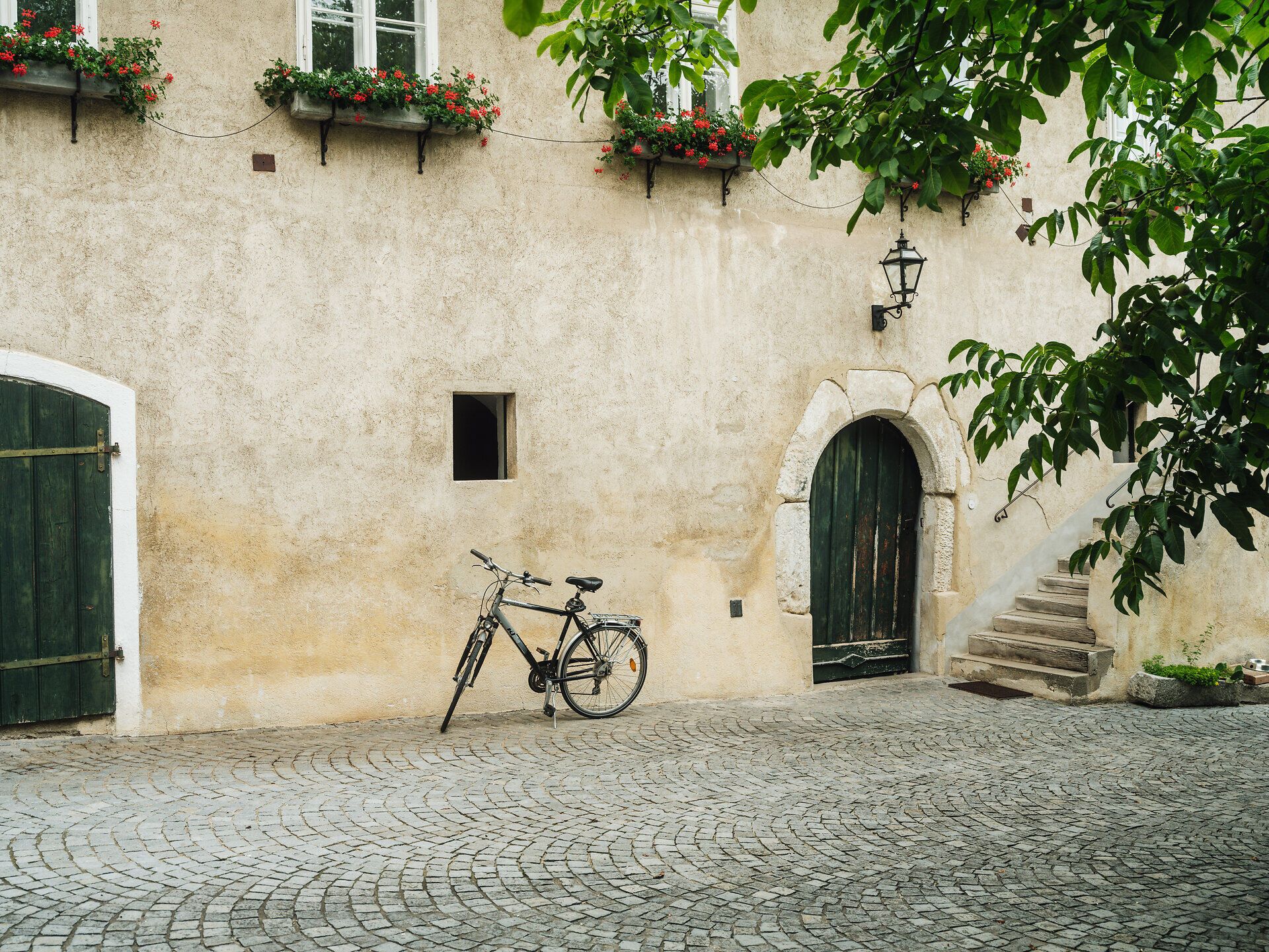Fahrrad, steht vor einem alten Haus, an den Fenstern sind Blumenkisterl, alte grüne Türen und Pflastersteinboden.