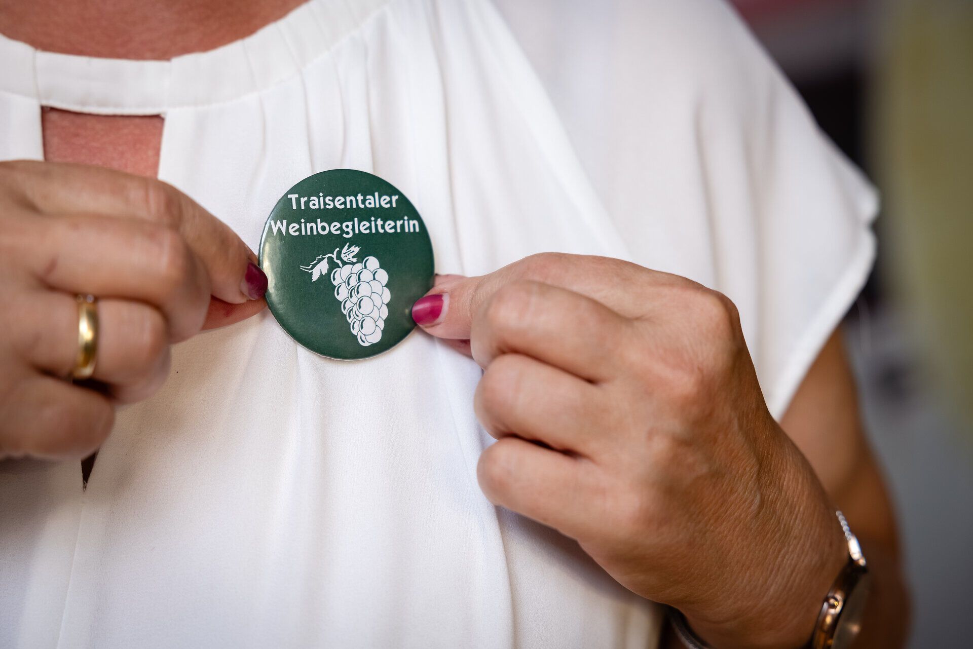 Blonde Frau von den Traisentaler Weinbegleitern lächelt hält den angesteckten, runden Pin mit dem Schriftzug Traisentaler Weinbegleiterin in die Kamera.